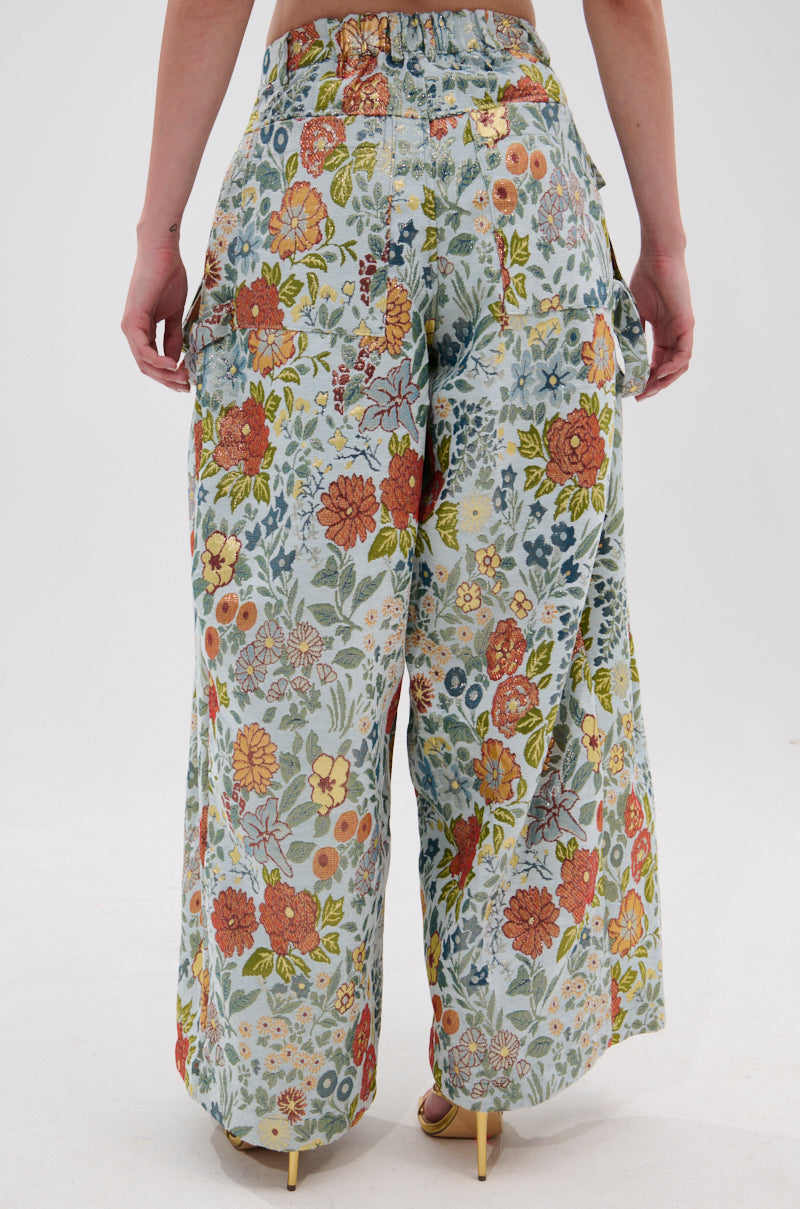 A person seen from the back wears MARTHA FLORAL CARGO PANTS—high-waisted, wide-leg with orange, yellow, and green tones, large pockets, and beige heeled shoes—creating a vintage feminine look. 3
