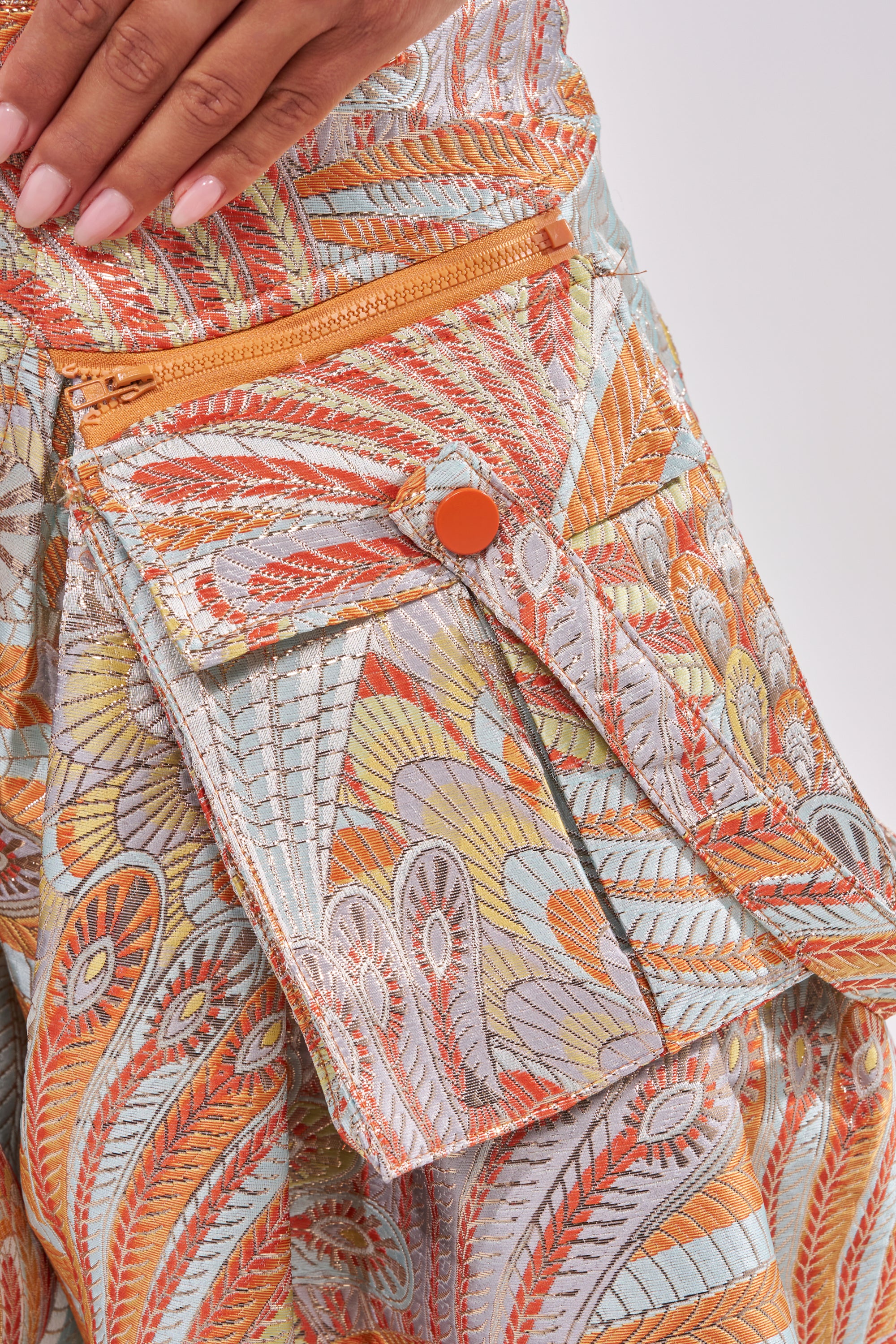 A close-up of a hand resting on MARTHA FLORAL TROUSER, showcasing blue wide-leg pants with a paisley-patterned pocket, orange button, matching zipper, and colorful embroidery in orange, yellow, and blue. 9