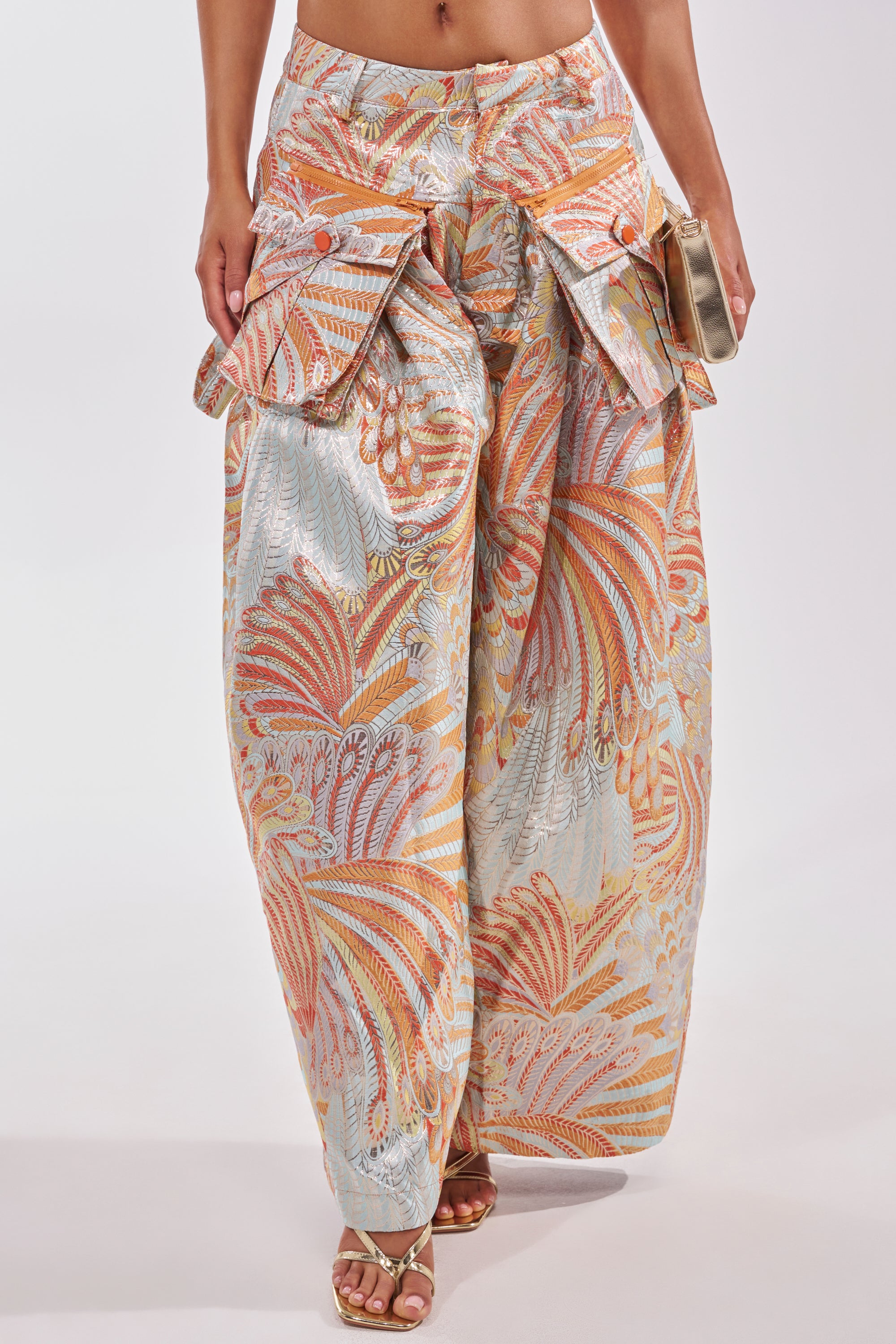 A woman models the MARTHA FLORAL TROUSER—wide-leg, high-waisted pants with a swirling orange, pink, and blue pattern, large front pockets, metallic details—styled with gold heels and a small clutch. 2
