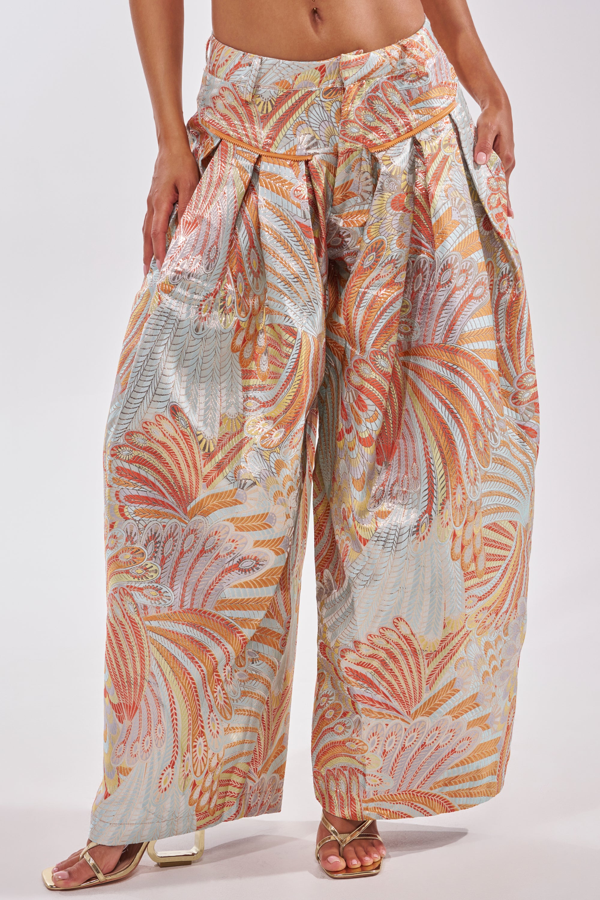 A person wears the MARTHA FLORAL TROUSER—blue wide-leg pants with a colorful abstract feather pattern in pastel orange, blue, and beige—paired with open-toe sandals, standing against a plain white background. 3