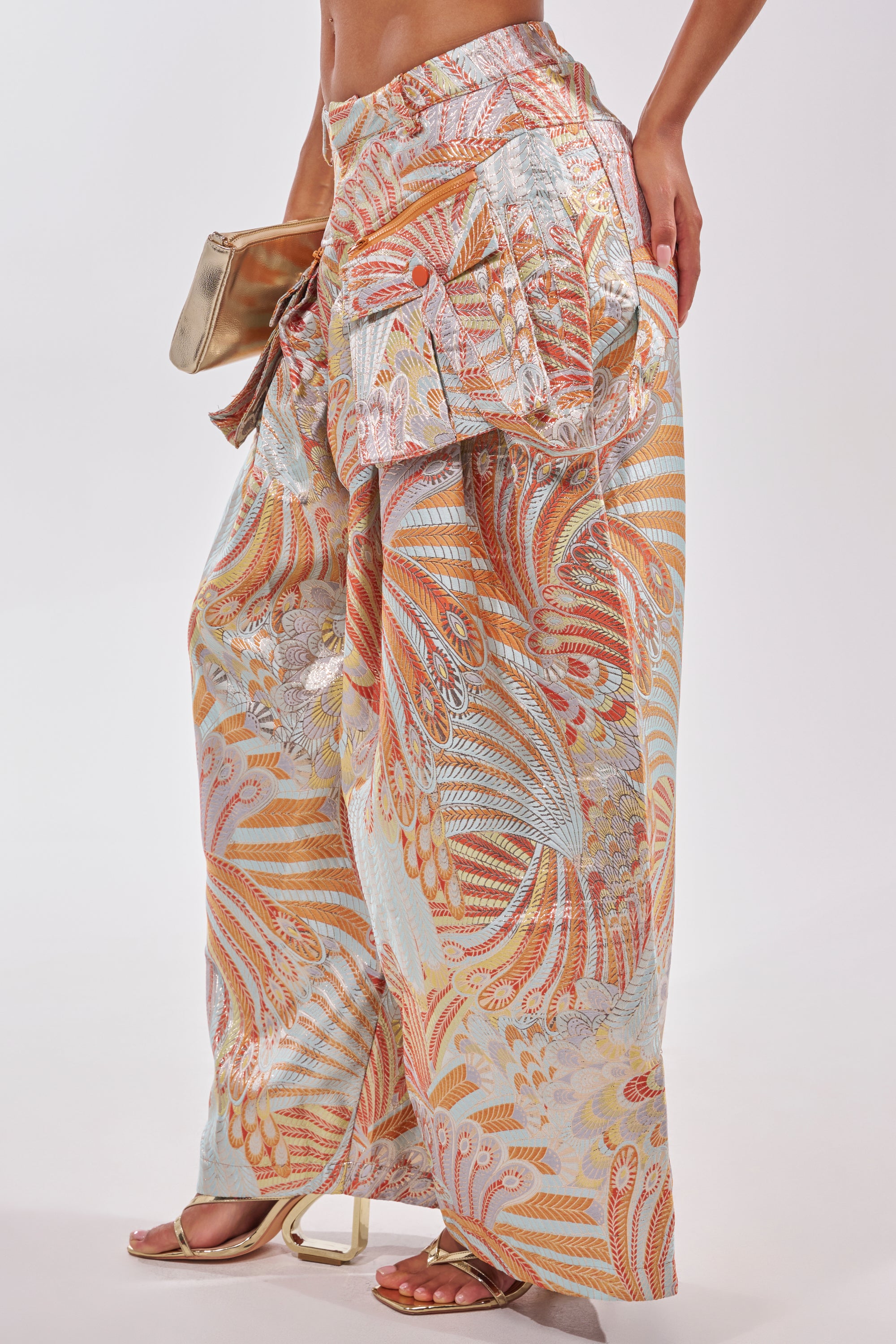 A person wearing the MARTHA FLORAL TROUSER—high-rise wide-leg pants with a vibrant orange, gold, and blue pattern—paired with metallic high-heeled sandals and holding a gold clutch. 4