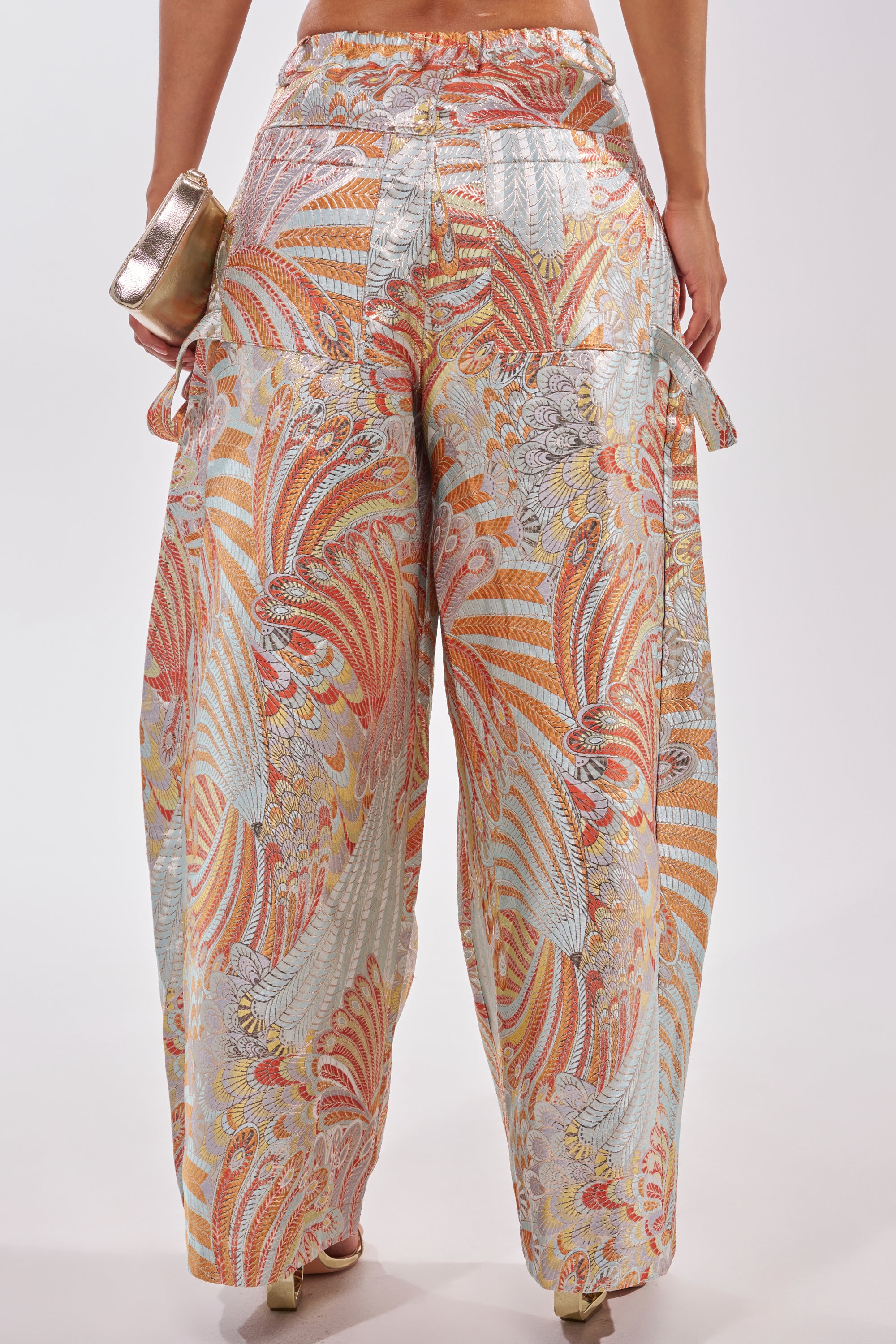 A person wears the MARTHA FLORAL TROUSER, featuring a high-rise wide leg and an intricate orange, gold, and light blue swirling floral pattern, holding a metallic handbag against a plain white background. 5