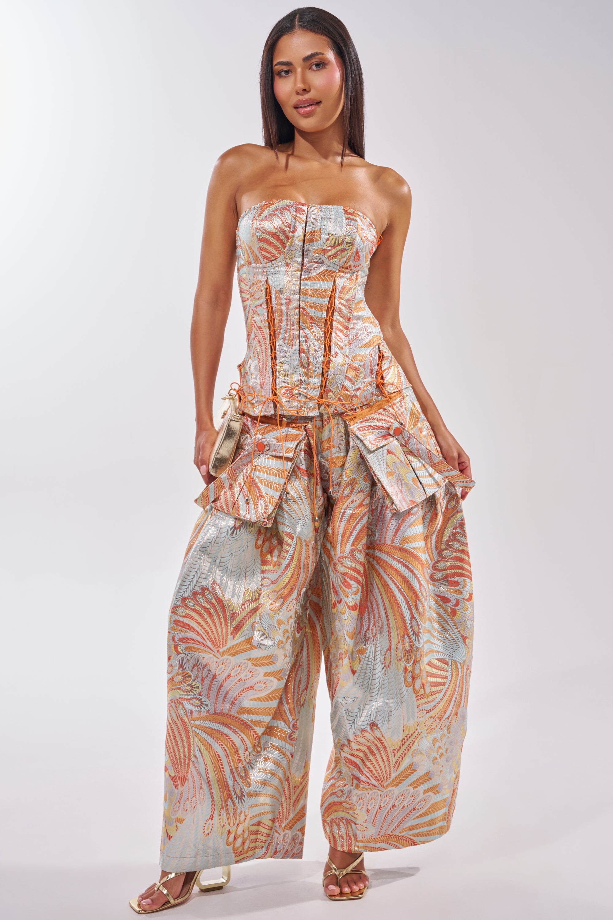 A woman wears the MARTHA FLORAL TROUSER, featuring high-rise wide leg pants in pastel orange, blue, and gold with a fitted, strapless bodice. She accessorizes with a metallic clutch and heeled sandals on a plain background. 6