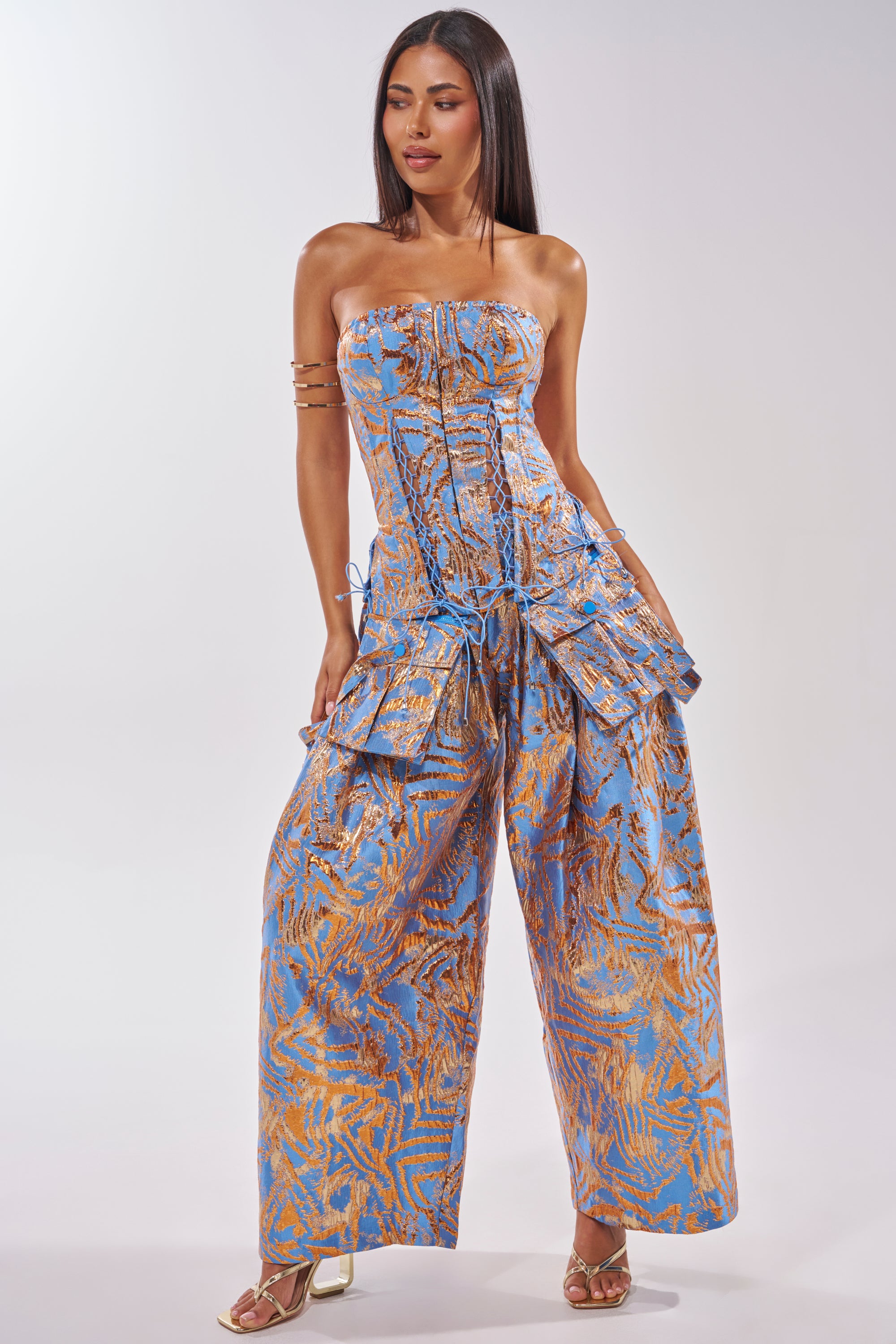 A woman wears the WAYS TO GO BROCADE TROUSER, a strapless blue jumpsuit with a high-rise waistband, gold leaf patterns, wide-leg trousers, and ruched details. She has straight dark hair, gold arm cuffs, and strappy heels against a plain background. 0