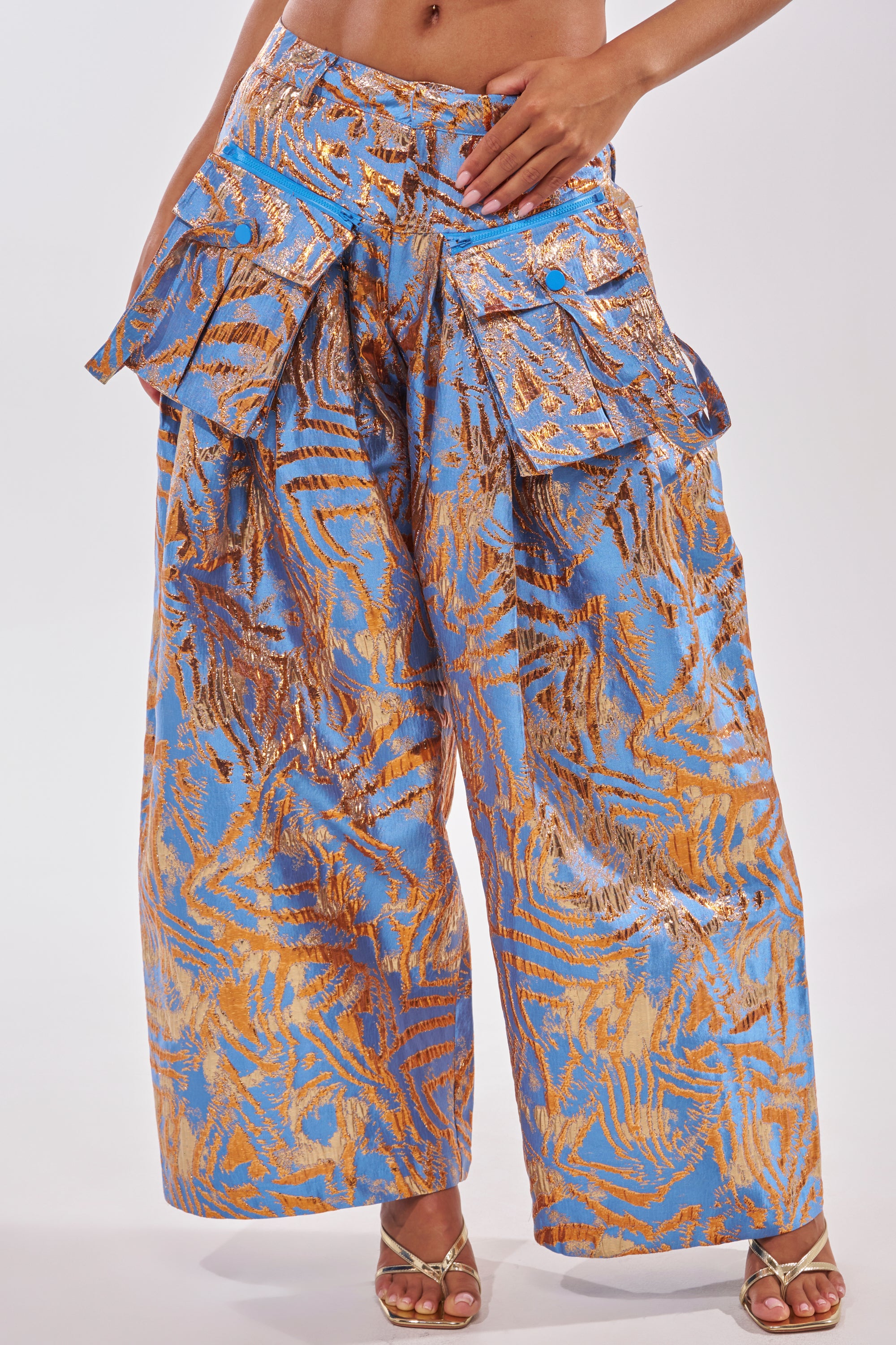 Wearing the WAYS TO GO BROCADE TROUSER, this look features shiny blue brocade trousers with a wide-leg silhouette, gold abstract pattern, large front pockets, and gold high-heeled sandals—perfect for standout cocktail party style. 1