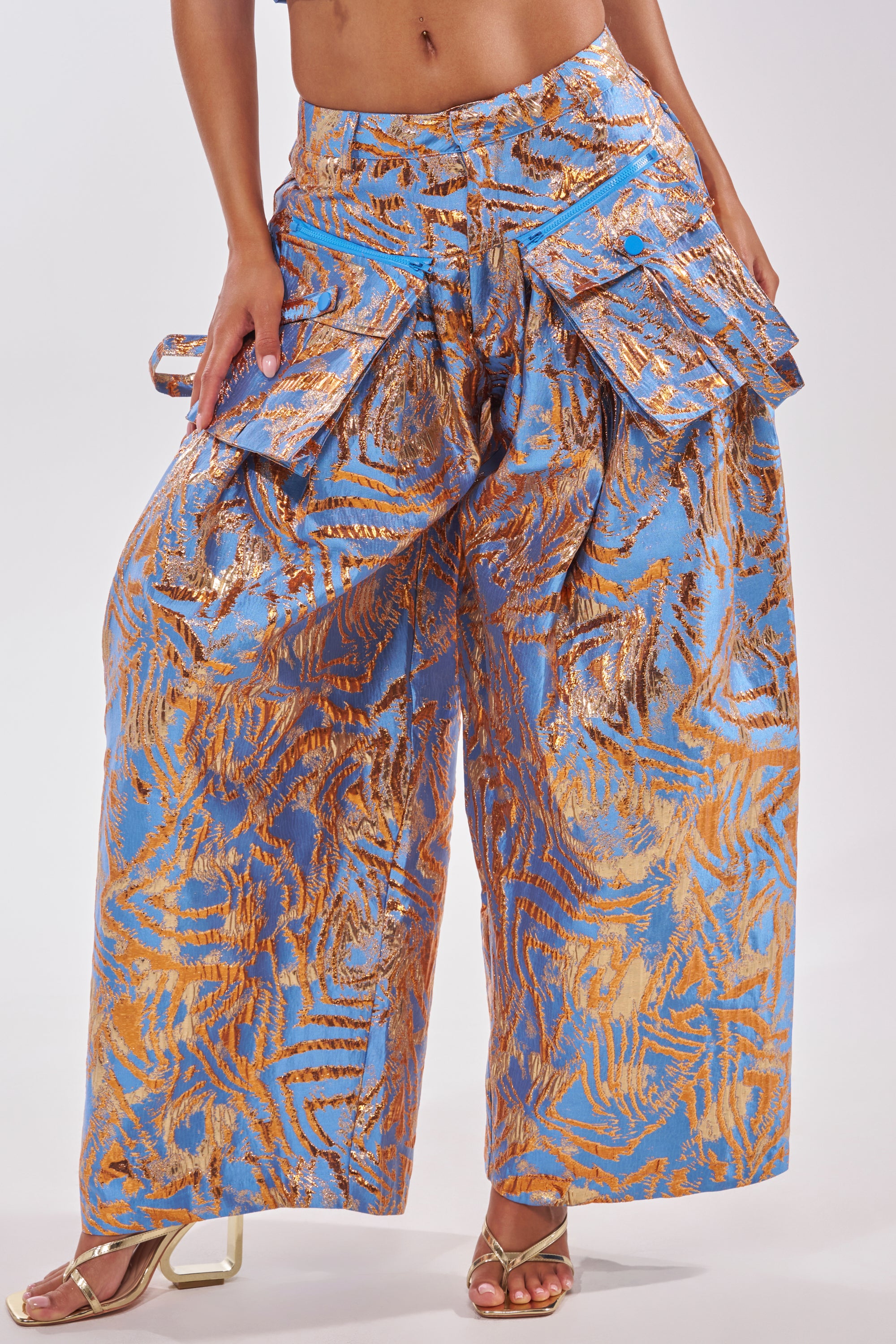 The WAYS TO GO BROCADE TROUSER features a wide-leg, high-waisted fit with large layered pockets and a matching belt. Styled with strappy heeled sandals, these blue trousers make a bold midriff-baring statement for cocktail parties. 2