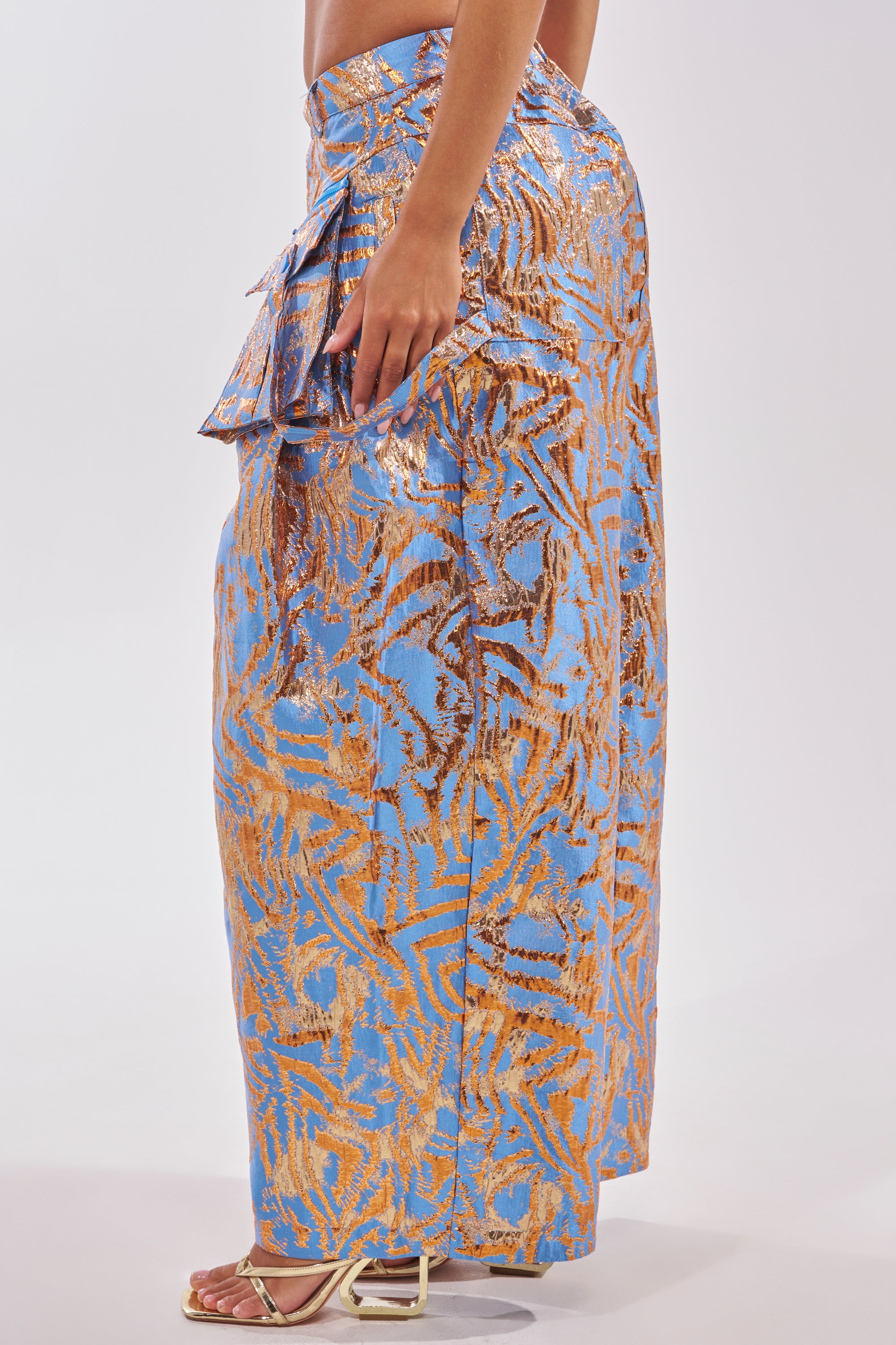 Model wearing the WAYS TO GO BROCADE TROUSER, featuring a metallic gold and blue pattern, side ruffle detail, and wide-leg silhouette with strappy sandals. Side view on a plain white background. 3