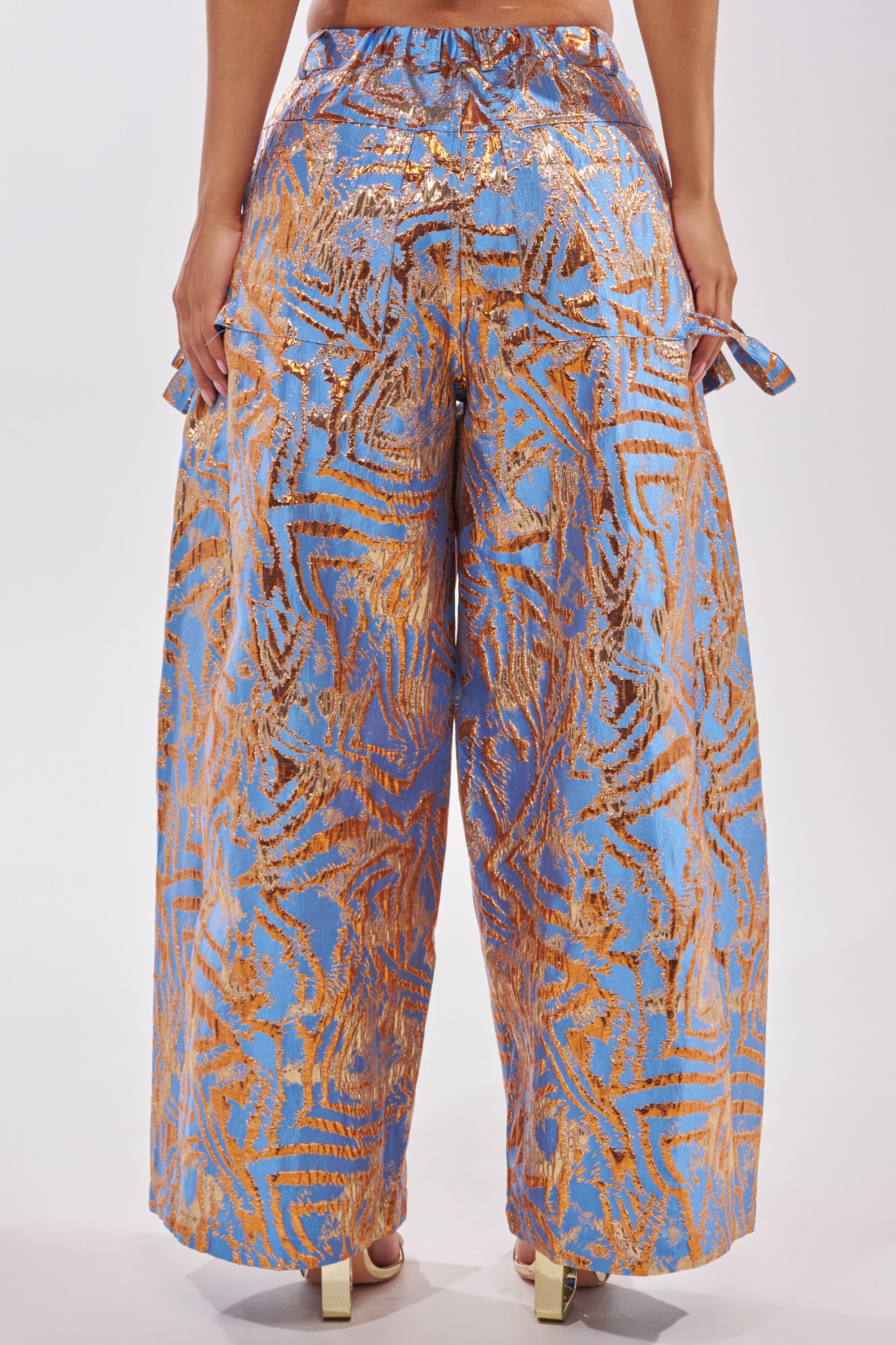 A person is seen from the back wearing WAYS TO GO BROCADE TROUSER: high-waisted, wide-leg blue trousers with a metallic gold abstract pattern, paired with light-colored heels—a stunning choice for any cocktail party. 4