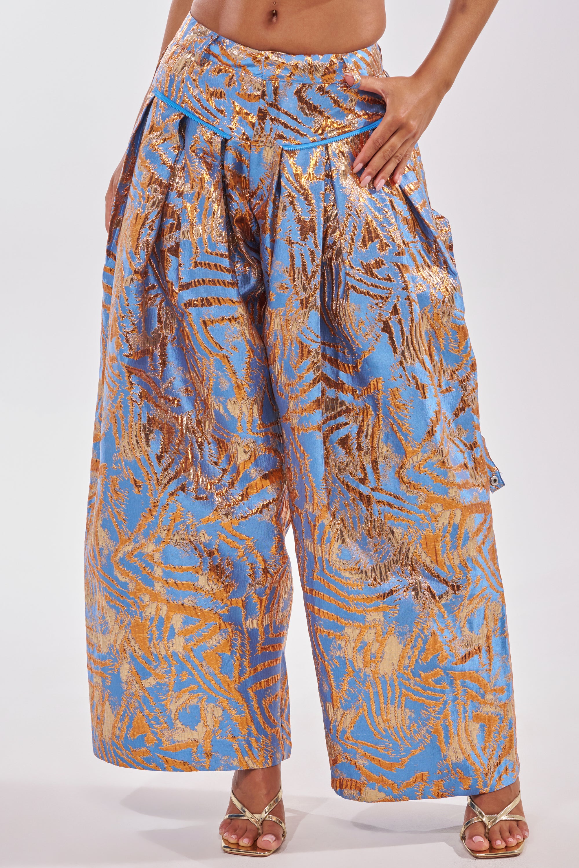 A person wears WAYS TO GO BROCADE TROUSER—wide-leg, high-waisted blue pants with a shiny gold abstract pattern—paired with open-toe heeled sandals. Only the lower half of the body is visible. 5