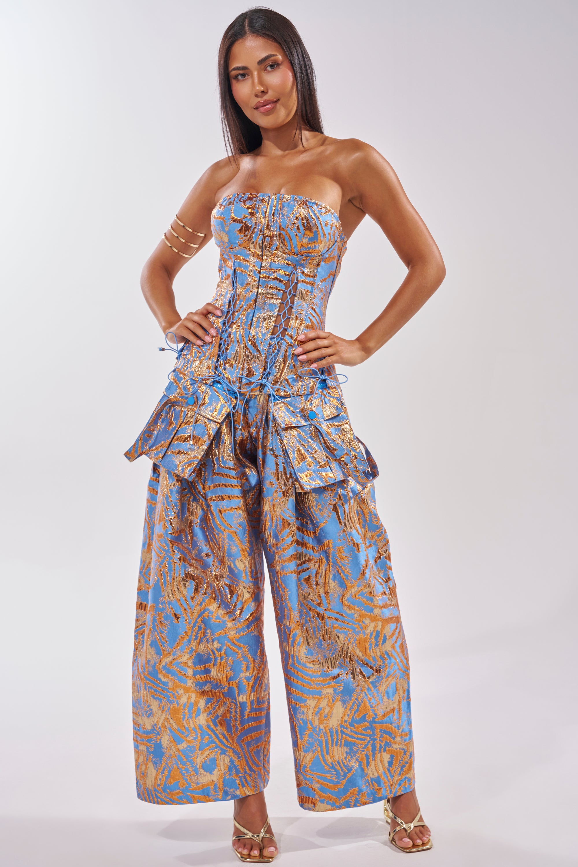 A woman stands confidently in WAYS TO GO BROCADE TROUSER, styled as a strapless jumpsuit with gold patterns, layered peplum details, gold heels, and a gold arm cuff against a plain light background. 7