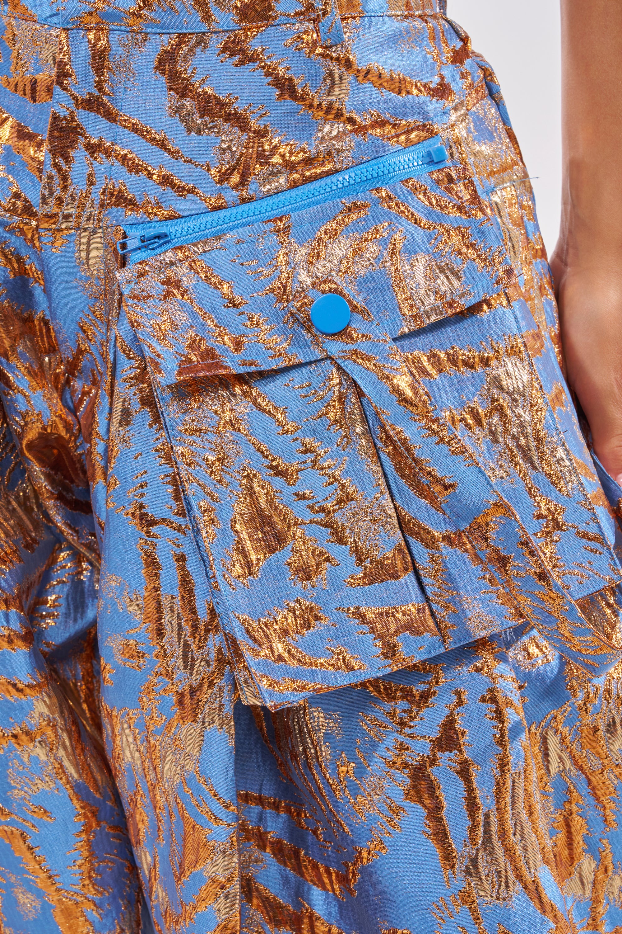 Close-up of WAYS TO GO BROCADE TROUSER: blue fabric with shiny gold abstract patterns, a pocket with blue button and matching zipper. A person's arm appears on the right, accentuating these chic cocktail party pants. 8