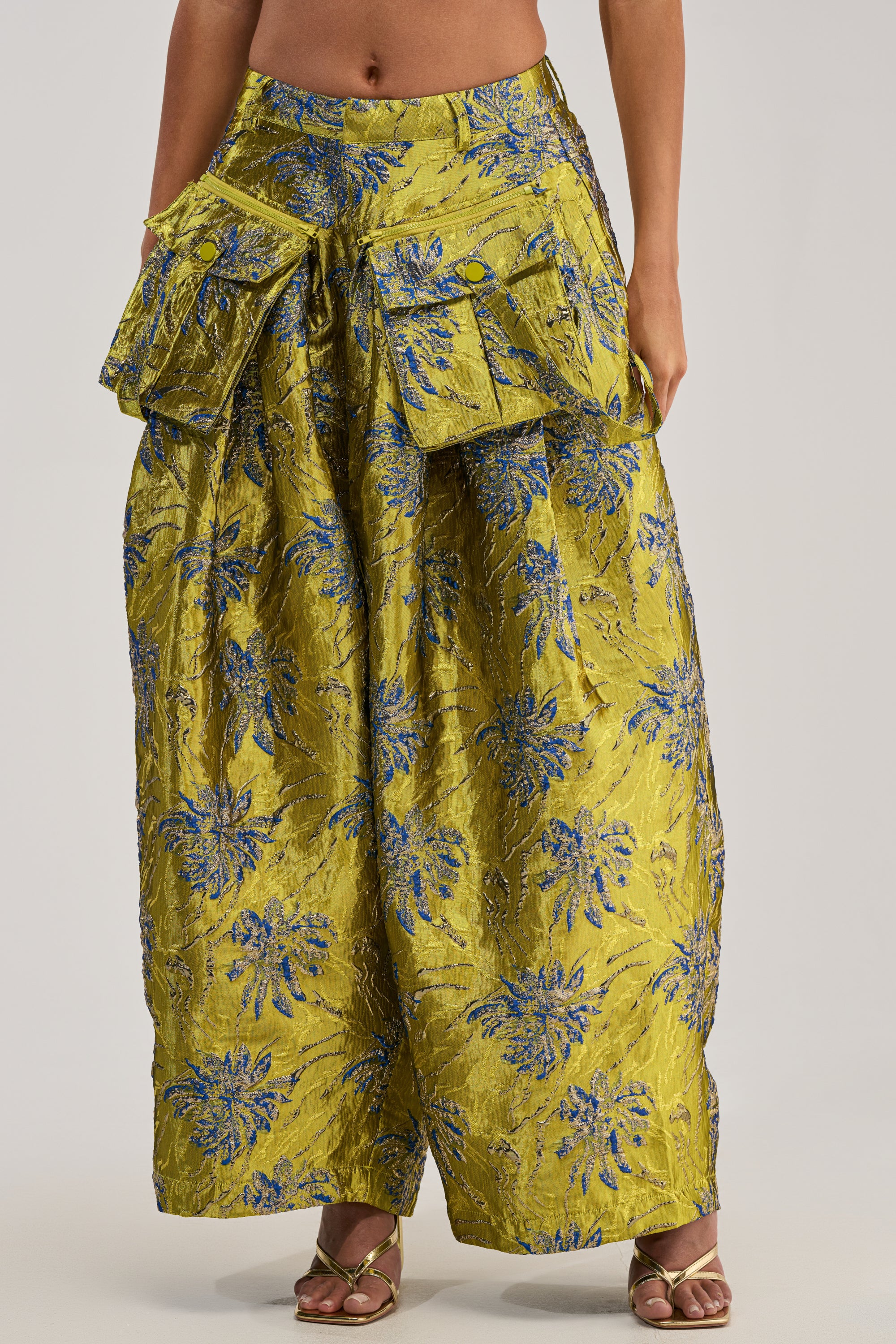 A person wears the CRAZY FOR YOU BROCADE TROUSER—wide-legged gold pants with blue floral patterns, two large hip flap pockets—and gold open-toed heels. Upper body is not visible.