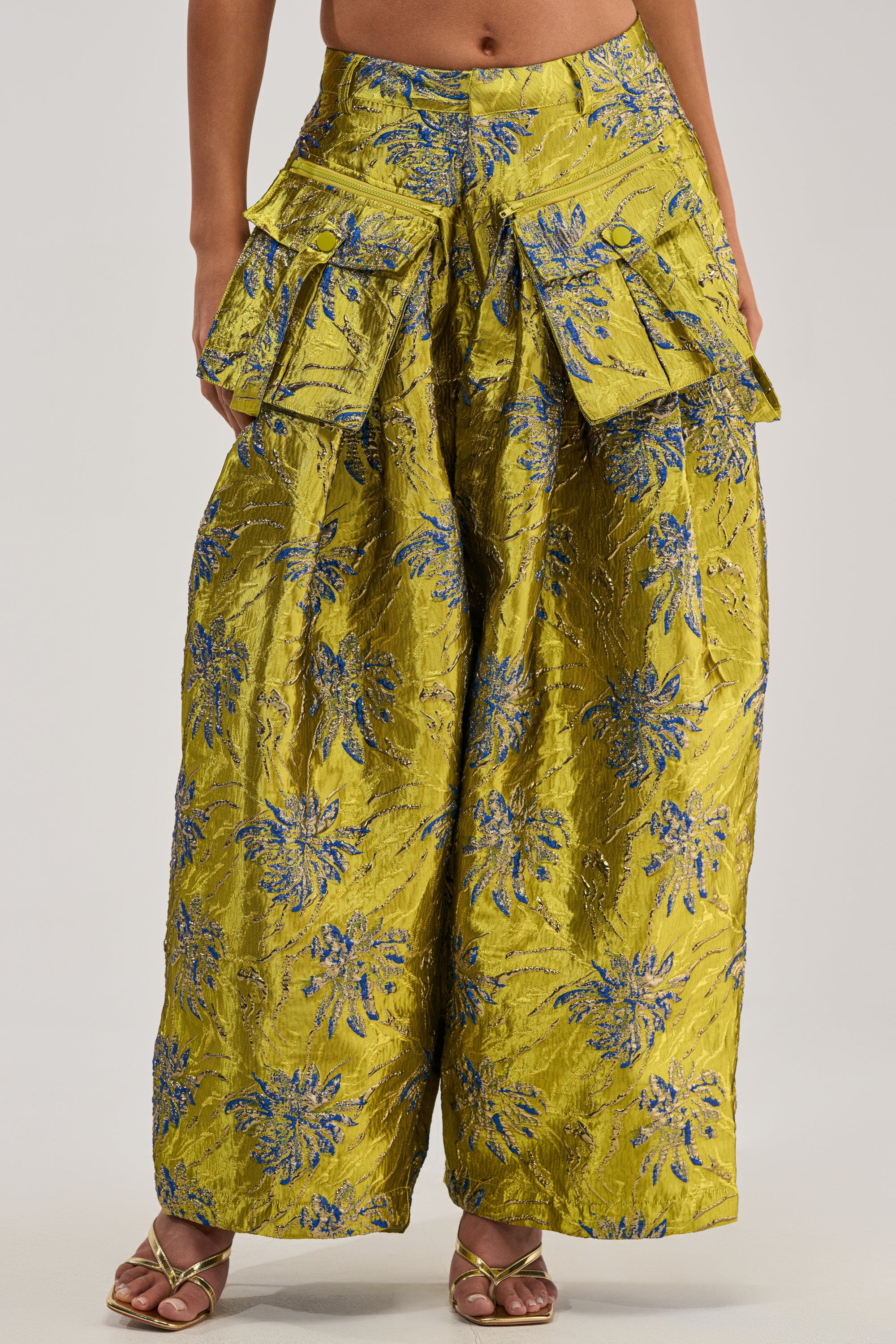 A person wearing the CRAZY FOR YOU BROCADE TROUSER, featuring a wide-leg silhouette, shiny yellow-green fabric with blue floral patterns, large flap pockets, and paired with heeled sandals.