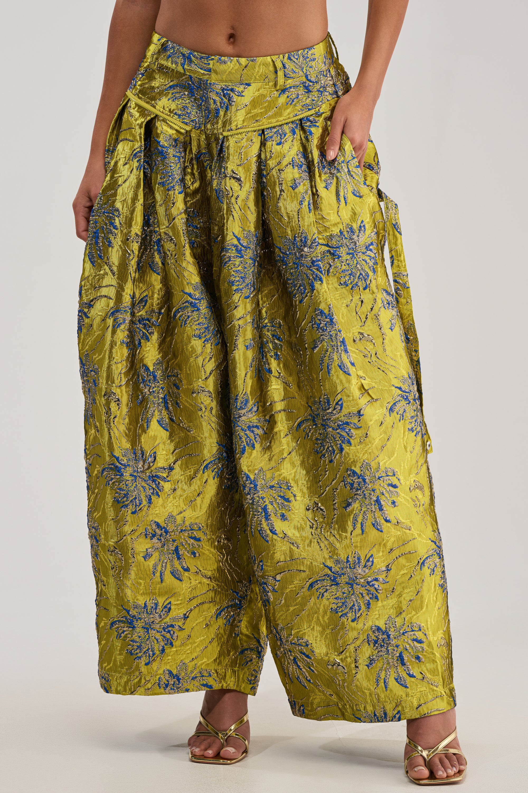 Only the lower torso and legs are visible, wearing the CRAZY FOR YOU BROCADE TROUSER—high-waisted, wide-legged brocade trousers in metallic yellow-green with blue floral patterns and pleats—paired with gold open-toe heels.