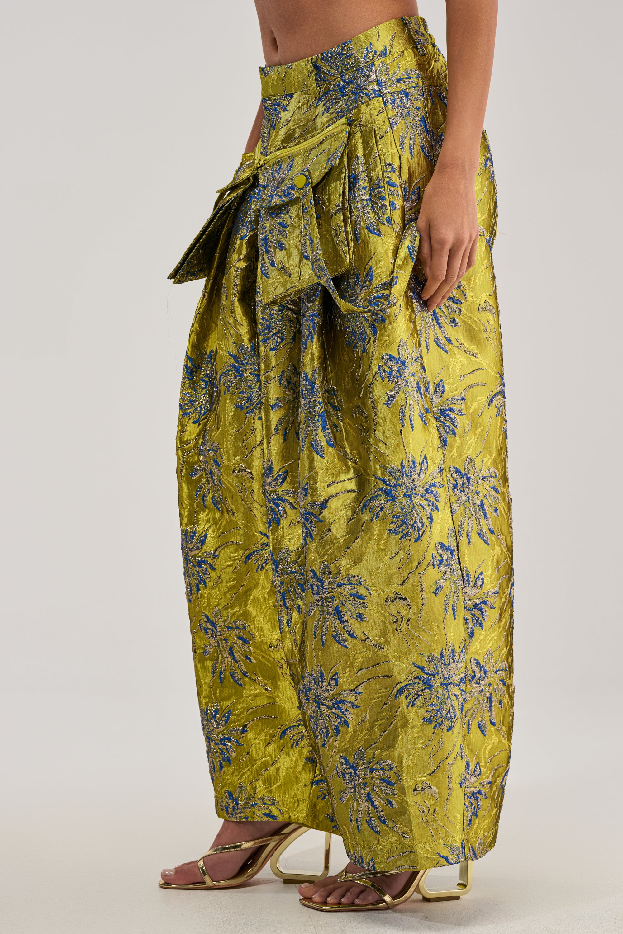 A person models the CRAZY FOR YOU BROCADE TROUSER—a long, shiny yellow pant with blue floral patterns, a large waist bow, and an elegant wide-leg silhouette. Gold high-heeled sandals are worn; the upper body is mostly out of frame.