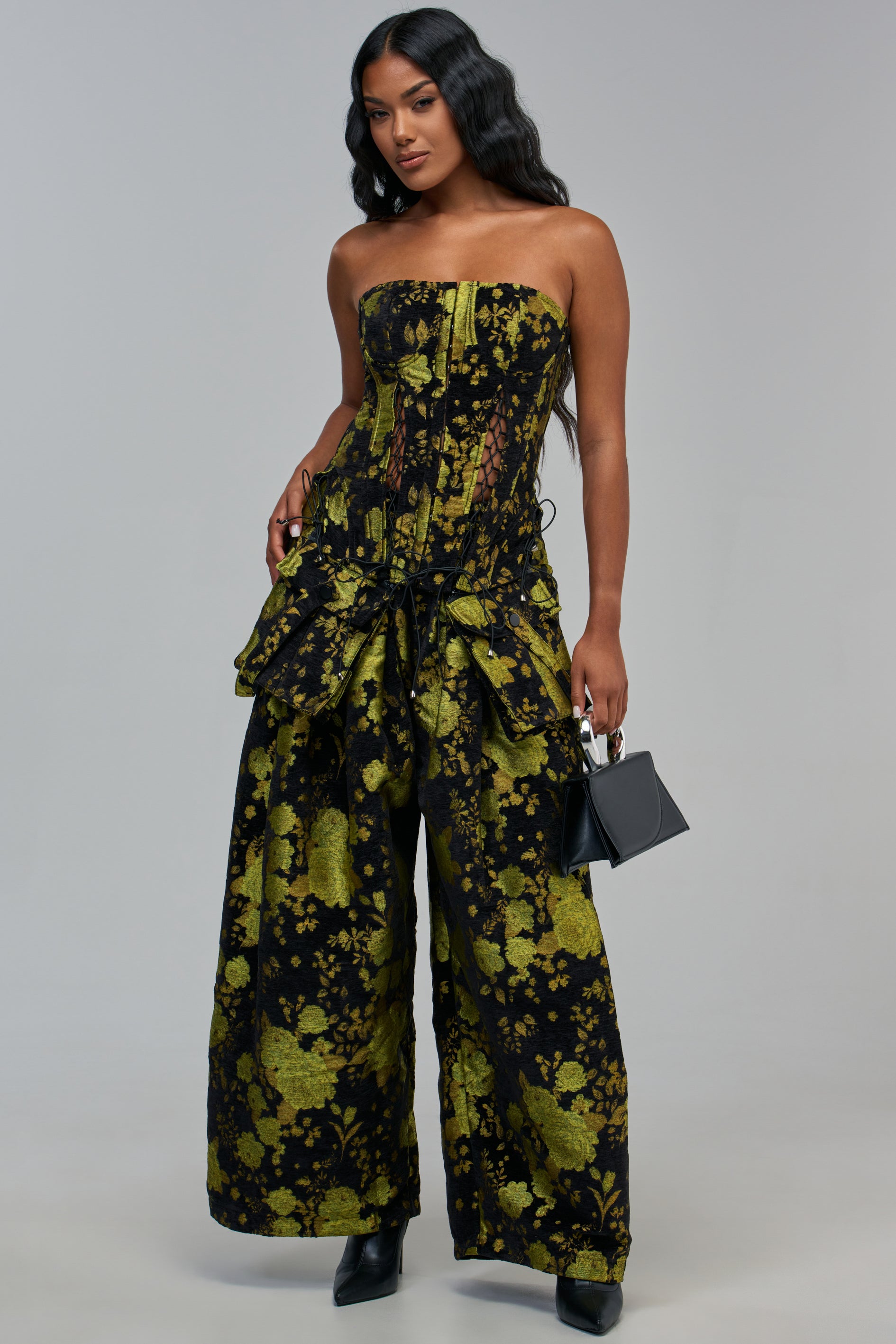 A model wears a black and lime green floral tapestry corset top with matching WAYS TO GO GREEN TAPESTRY TROUSER high-rise wide-leg pants, holding a small black handbag.