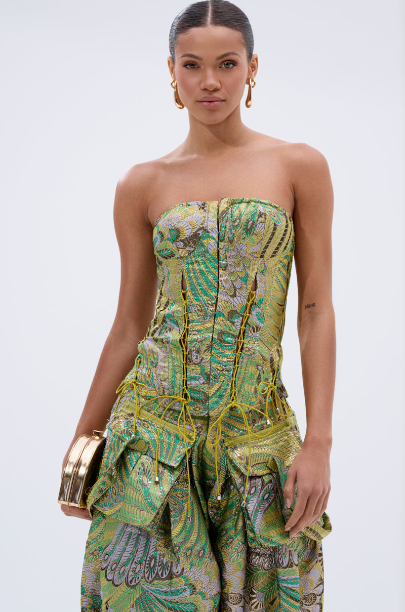 A woman wears the PEACOCK CORSET, a strapless dress with lace-up details, paired with gold hoop earrings and a gold clutch. Her hair is pulled back as she stands against a light background.