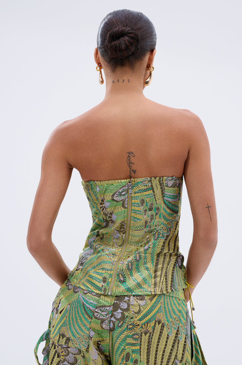 A woman with her back to the camera wears the PEACOCK CORSET—a strapless, green and gold top—with matching pants. Her hair is in a neat bun, and tattoos are visible on her neck, upper back, and left arm.