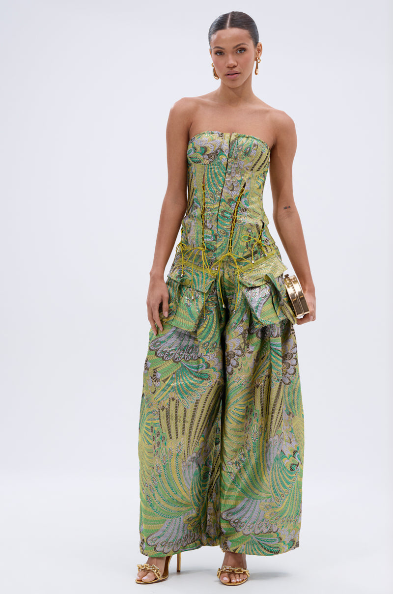A woman faces forward wearing the PEACOCK CORSET, a strapless brocade jumpsuit with green and gold patterns, wide-leg pants, gold heels, hoop earrings, and holding a gold clutch against a plain light background.