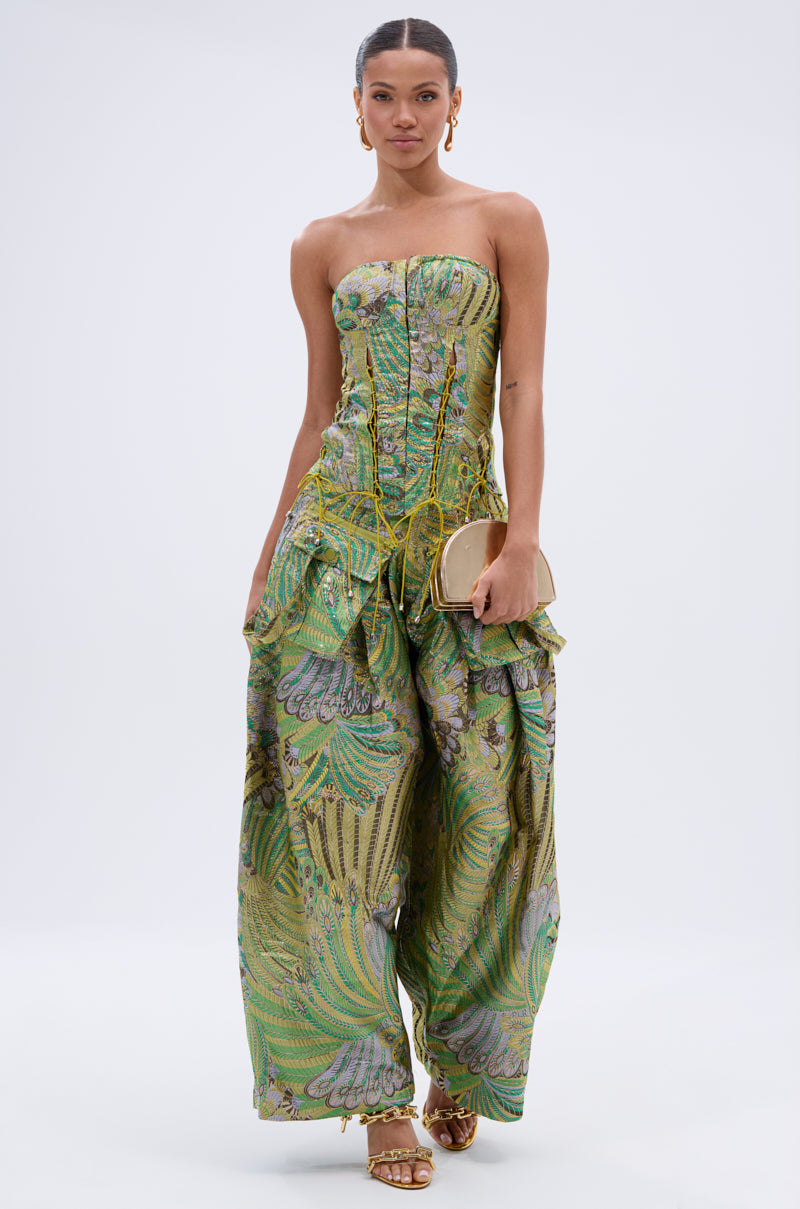 A woman wears the PEACOCK CORSET, a strapless green and gold brocade jumpsuit with wide-leg pants, holding a small gold purse against a plain white background.