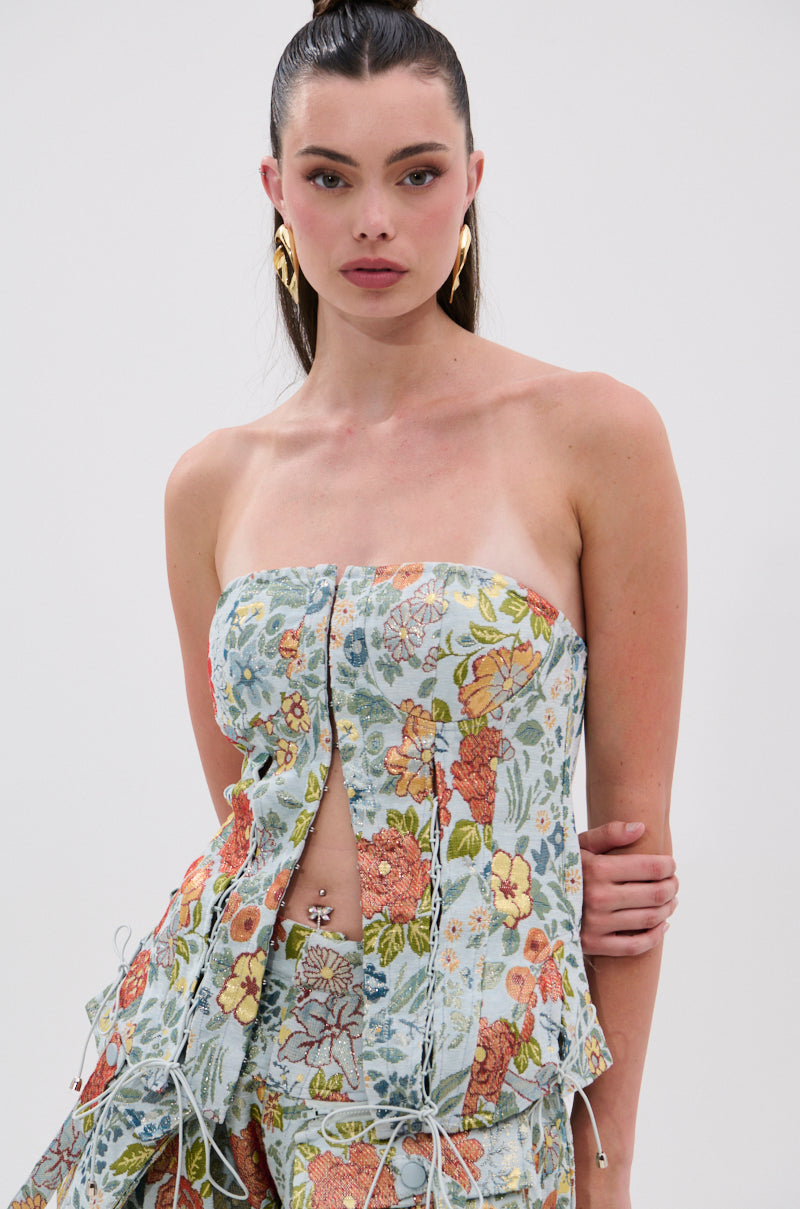 A woman with long dark hair in a high ponytail wears the MARTHA FLORAL CORSET and matching pants, accessorized with gold earrings, posing against a plain light background.