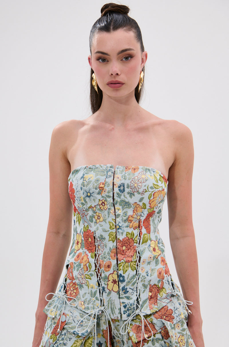 A woman with long dark hair in a high bun wears the MARTHA FLORAL CORSET, a vintage-inspired strapless dress with a fitted bodice and gold earrings, standing against a plain white background.