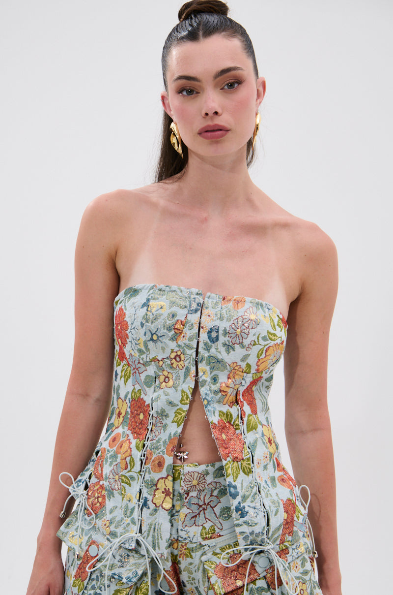 Woman with long dark hair in a high ponytail, gold hoop earrings, and the vintage-inspired strapless MARTHA FLORAL CORSET in light blue floral print with ties and matching pants, standing against a plain white background.
