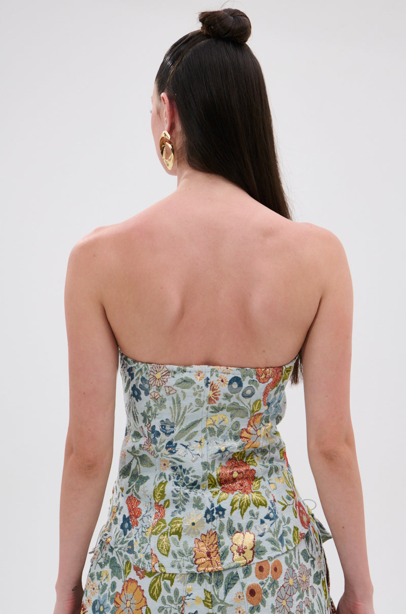 A woman with long dark hair in a topknot is seen from the back wearing the vintage-inspired MARTHA FLORAL CORSET, which features a colorful floral pattern. She accessorizes with large gold hoop earrings against a plain light background.