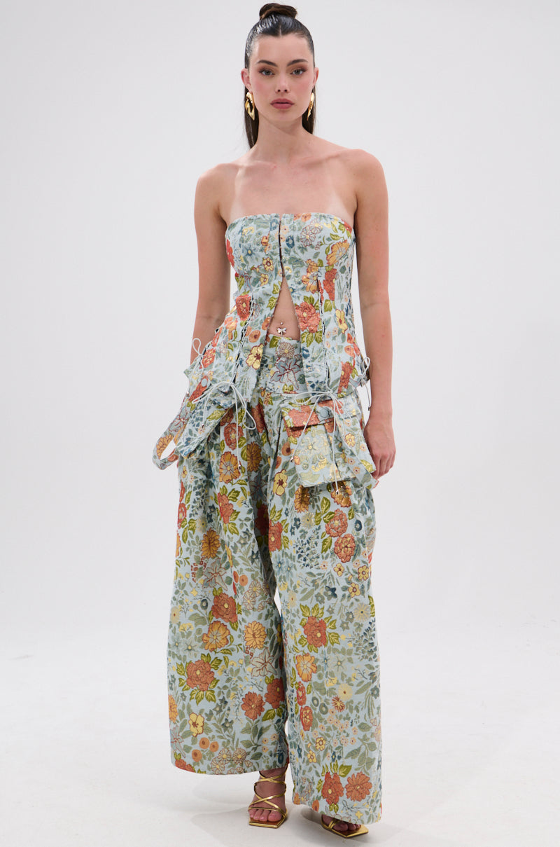 A woman stands against a white background in the MARTHA FLORAL CORSET, a strapless top with front cutouts and matching wide-leg pants, both adorned with vibrant orange, yellow, and green flowers. She completes the look with heels and gold hoop earrings.