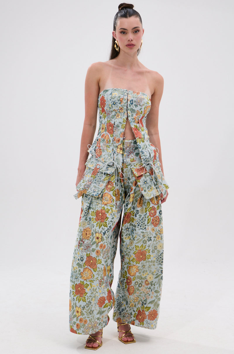 A woman wears the MARTHA FLORAL CORSET, a vintage-inspired outfit with cutouts, ruffled sides, and buttoned top paired with matching wide-leg pants. She accessorizes with gold earrings and a high ponytail against a plain background.