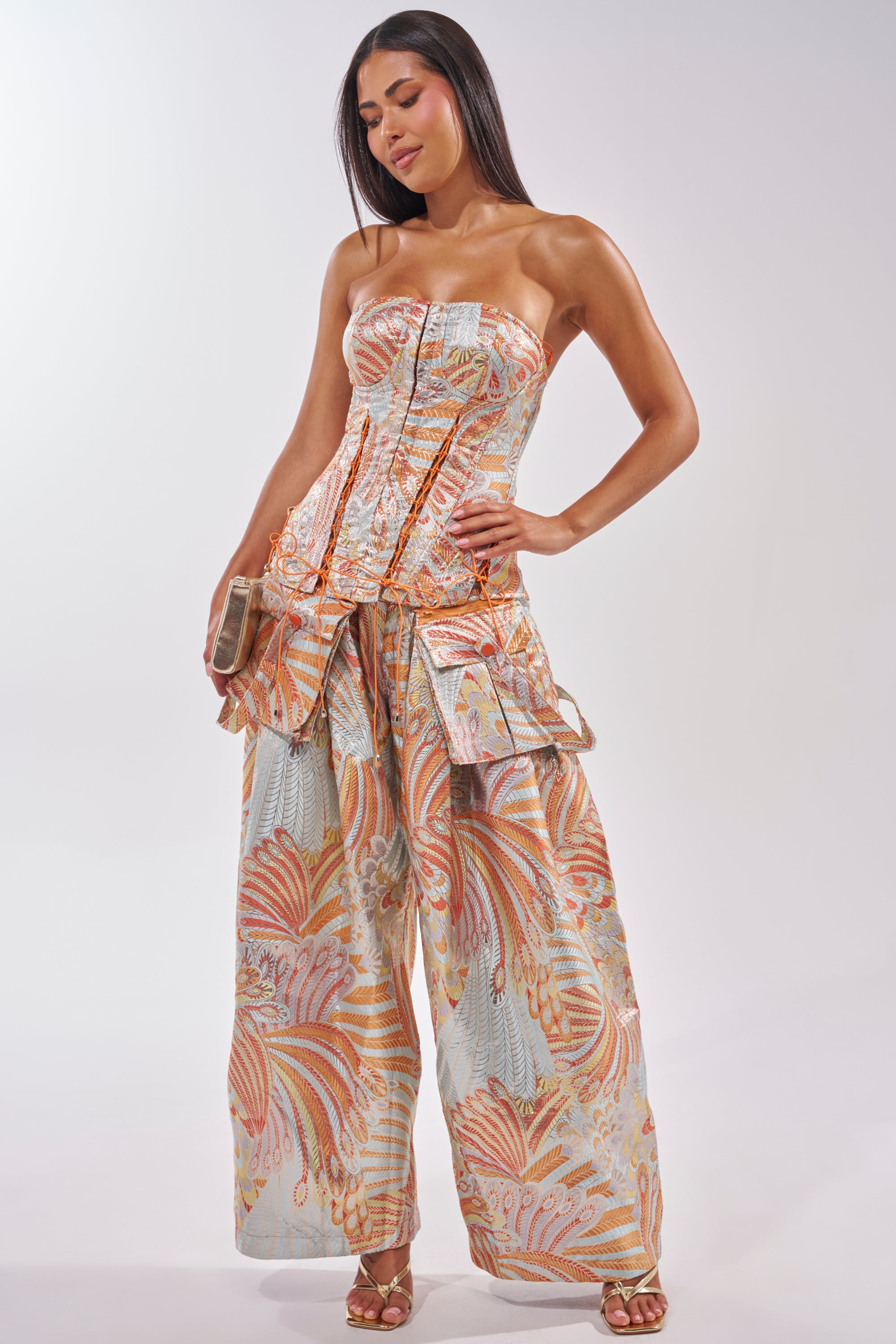 A woman poses confidently in the MARTHA FLORAL CORSET and wide-leg pants with a pastel geometric print. She accessorizes with a small clutch, strappy heels, and wears her straight hair down, creating a chic look. 4