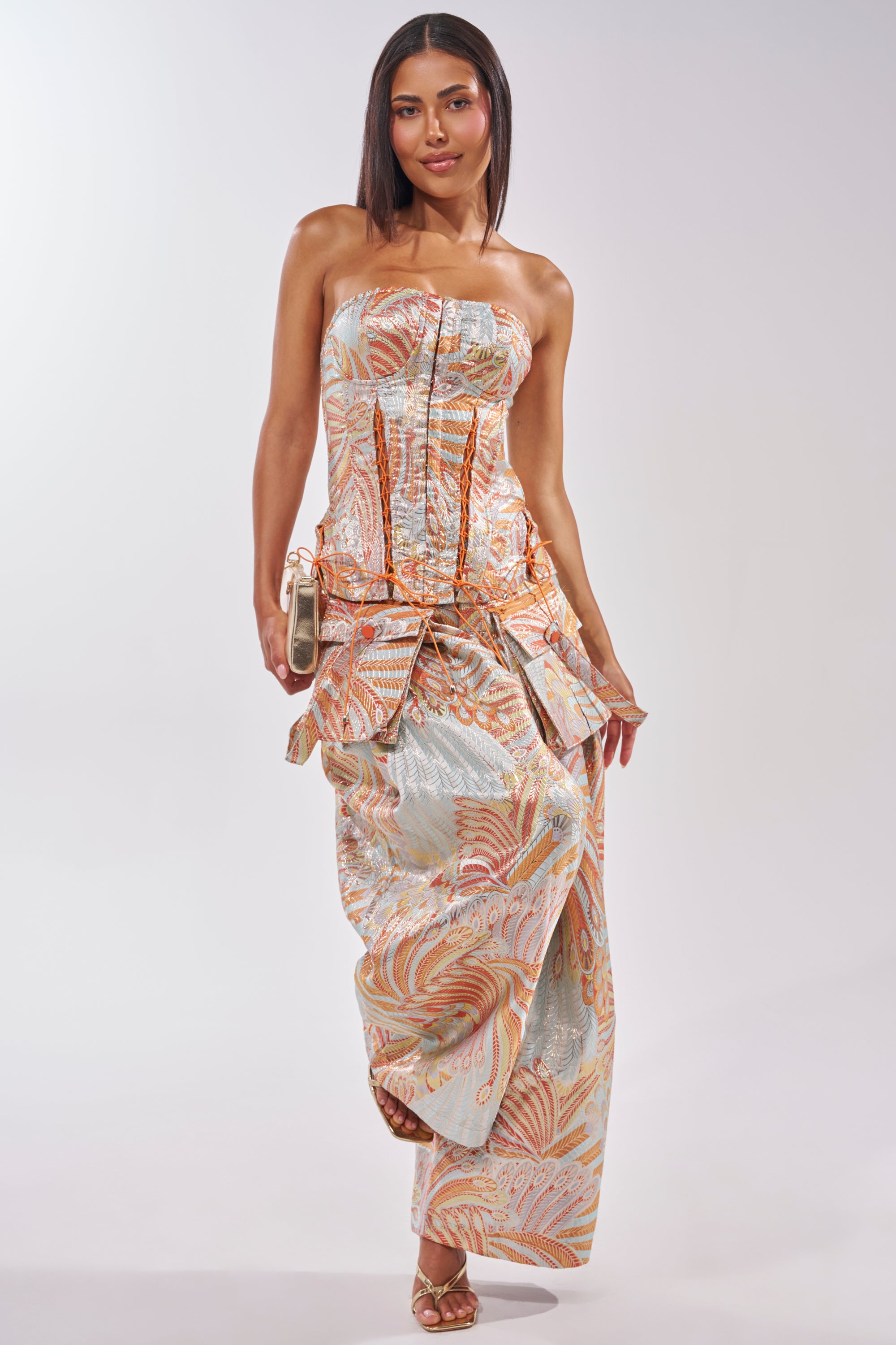 A woman confidently poses in the MARTHA FLORAL CORSET, a strapless dress with a structured bodice, draped skirt, and pastel floral print. She carries a small handbag and wears strappy heels against a plain light background. 6