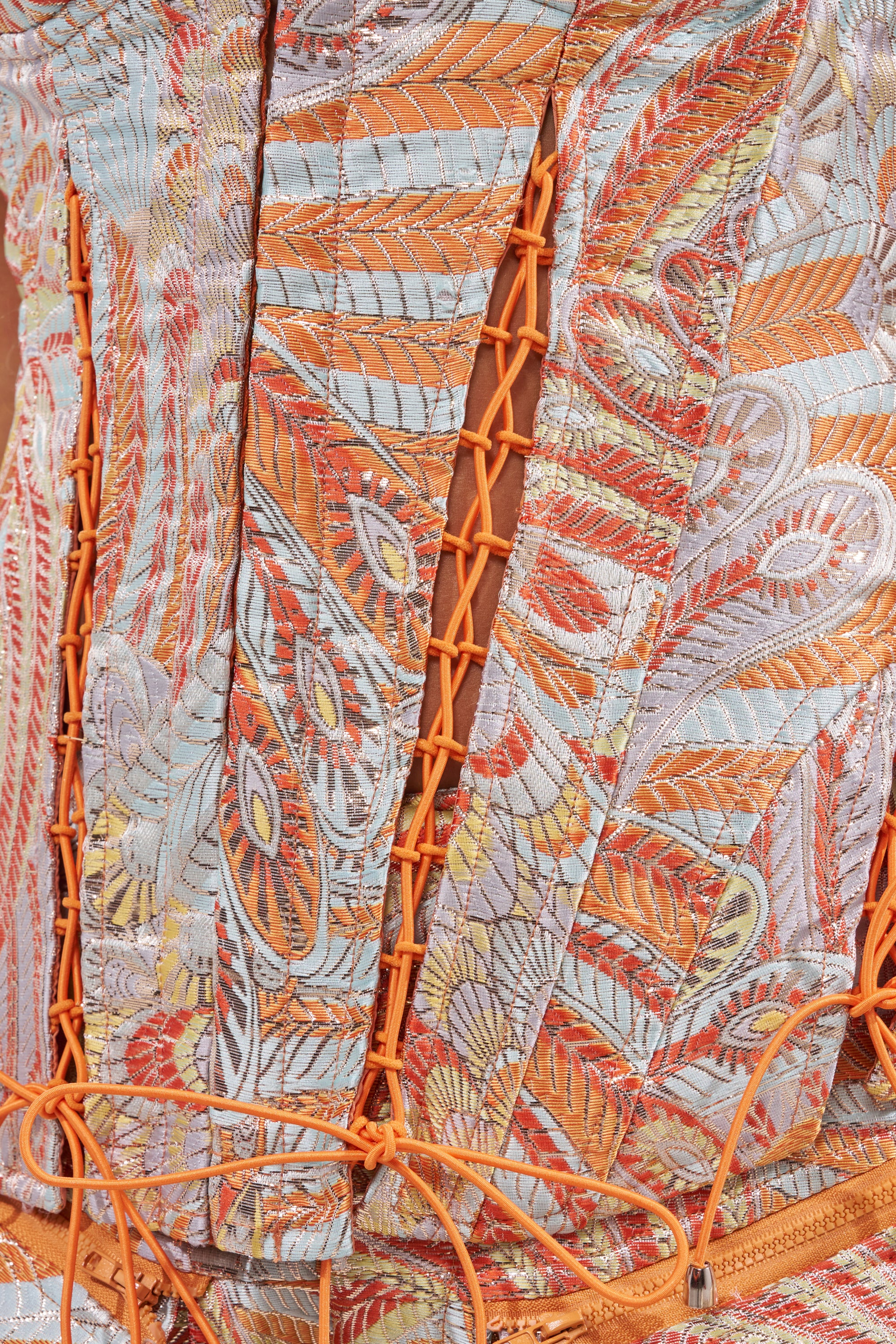 Close-up of the MARTHA FLORAL CORSET, showcasing colorful embroidered leaves and peacock feathers in orange, gold, and blue, with a lace-up panel featuring orange cord threaded through eyelets along the opening. 7