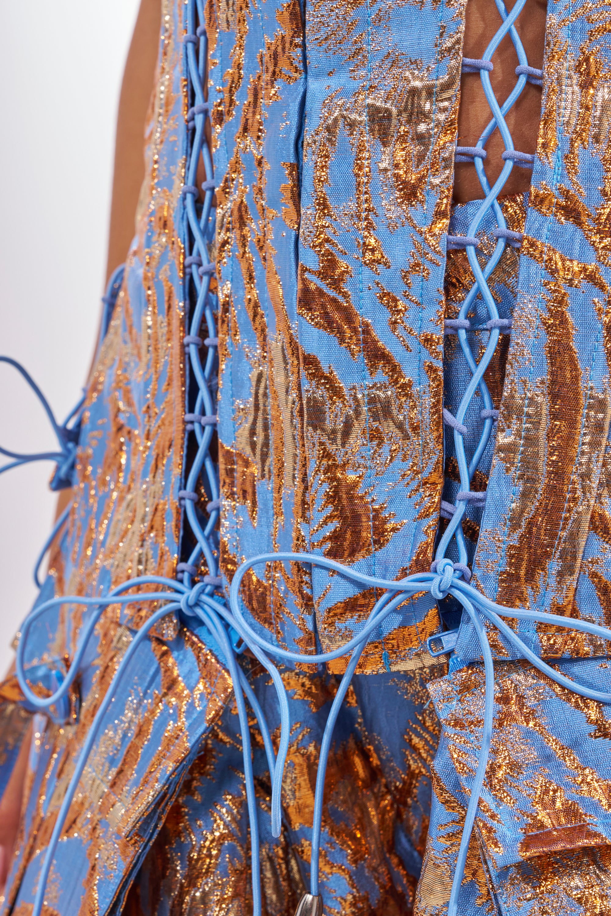 Close-up of the WAYS TO GO BROCADE CORSET in blue and metallic gold, featuring intricate lace-up paneling, bows, and a crisscross design that reveals skin beneath. The shimmering brocade fabric has reflective metallic detailing. 7