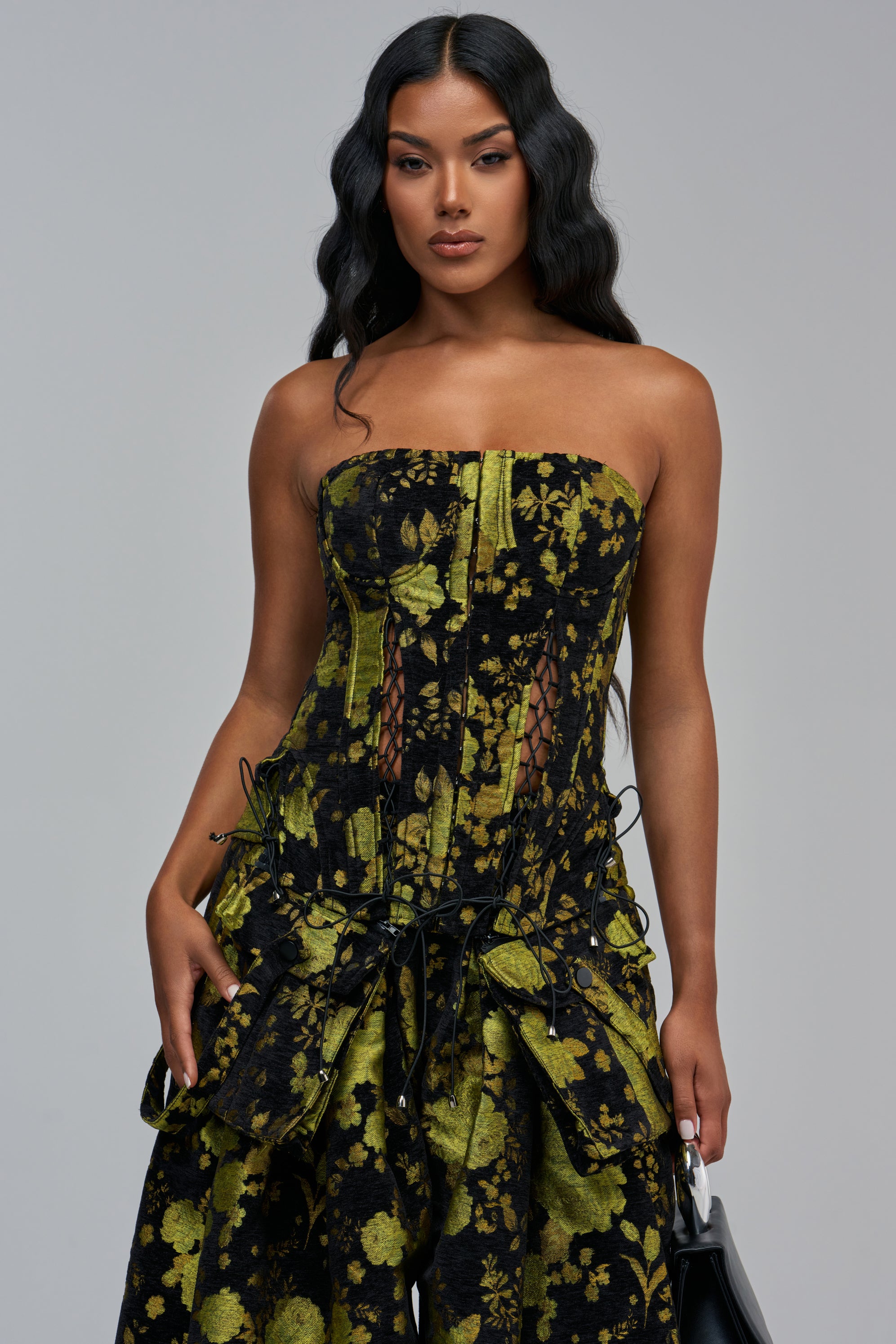 The WAYS TO GO GREEN TAPESTRY CORSET is a strapless floral top in a black and green tapestry pattern, featuring lace-up details at the midriff and pockets at the hem.