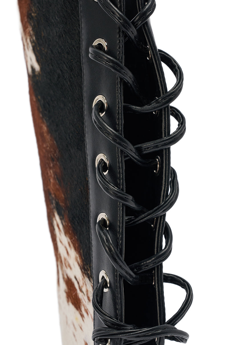 Close-up of black leather with silver eyelets and black laces on the AKIRA GOLD CALF HAIR LACE UP GLADIATOR STILETTO SANDAL, featuring brown and white calf hair, shown against a white background. 6