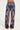 Someone is dressed in the DREAMING ABOUT YOU PANT, showcasing a high-rise waist and an intricate, symmetrical paisley design in blue, purple, and beige hues. These wide-leg pants are complemented by open-toed shoes against a plain off-white backdrop.