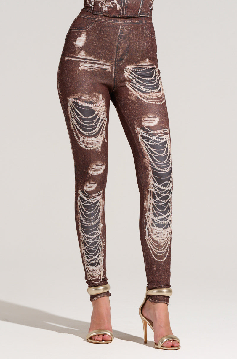 The woman flaunts high-waisted MILKY PANT leggings in brown, featuring distressed patterns and chain designs. The stretchy material mimics a mock denim look, complemented with gold high-heeled sandals for an elegant touch.