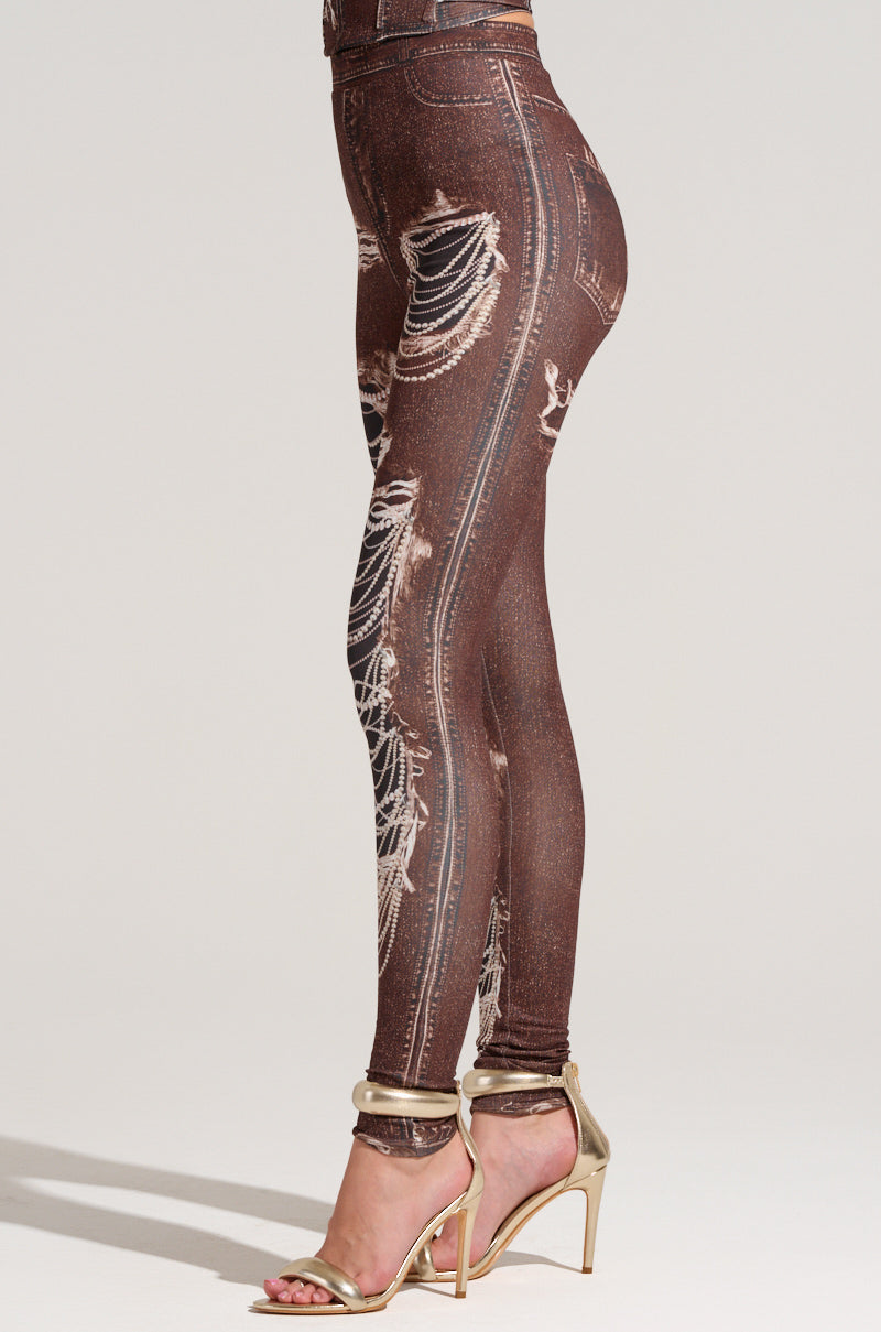 A person is wearing brown leggings featuring a mock denim, distressed jeans pattern design, known as the MILKY PANT. These stretchy fabrication leggings are paired with gold high-heeled sandals. The plain and neutral background enhances the chic modern look inspired by the AKIRA Label Milky Pant style.