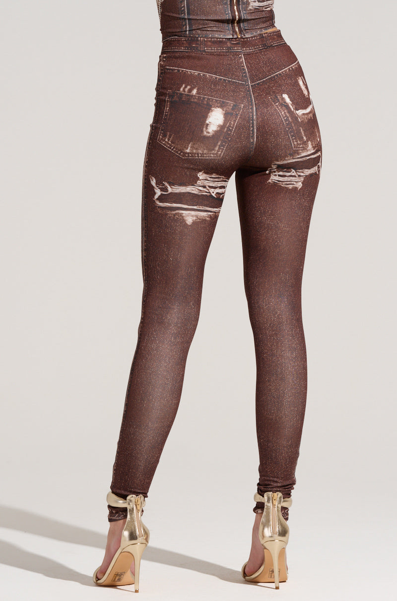 A person is dressed in distressed brown mock denim jeans, similar to the MILKY PANT, featuring a worn-out look with frayed details on both the pockets and legs. They are also wearing beige high-heeled sandals, all set against a plain white background.