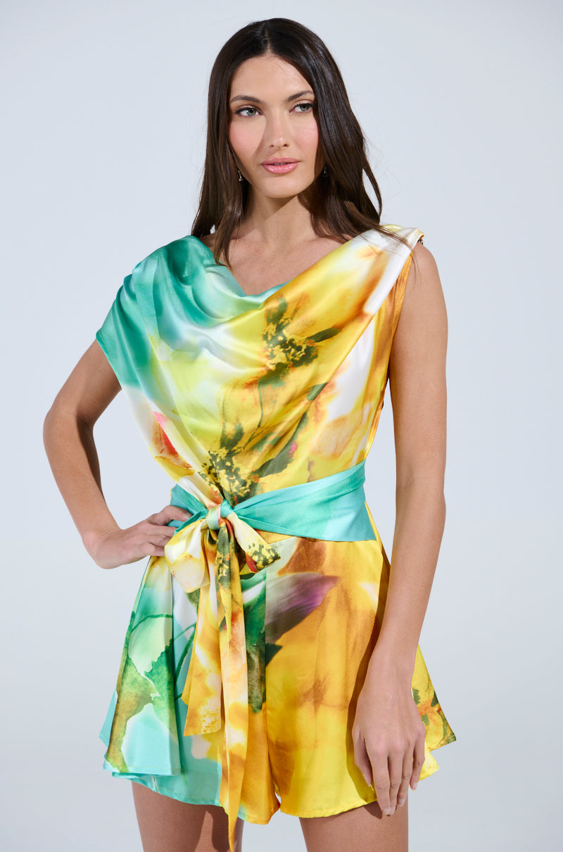 A woman with long dark hair wears the RALEIGH PRINTED SATIN ROMPER, featuring a watercolor floral print in green, yellow, and orange with a matching sash belt, standing against a plain light background.