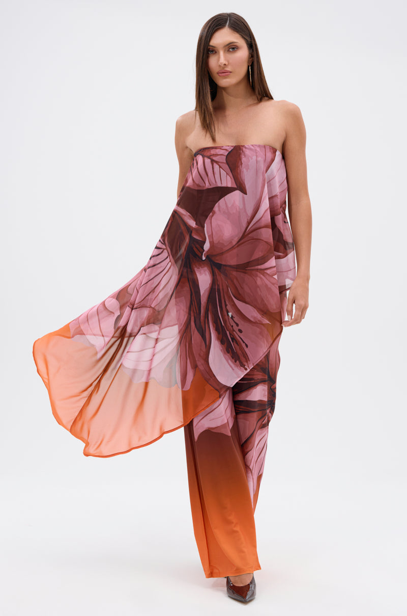 A woman with straight brown hair and a neutral expression poses against a plain backdrop in the ON A BEACH SOMEWHERE PRINTED JUMPSUIT IN ORANGE, a strapless, flowy jumpsuit with a tropical floral print and orange gradient—perfect for summer. 0