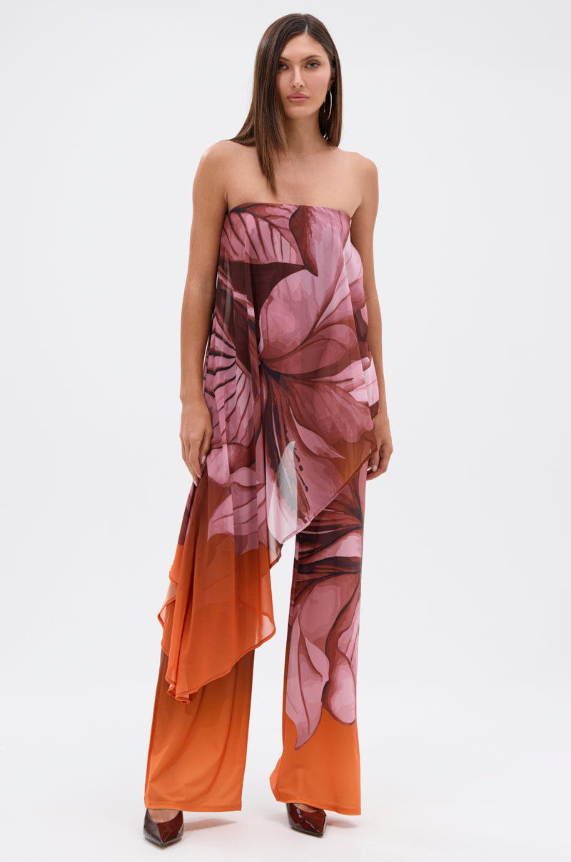 A woman wears the ON A BEACH SOMEWHERE PRINTED JUMPSUIT IN ORANGE, a strapless wide-leg jumpsuit with pink and orange tropical floral print, flowing asymmetrical overlay, and brown heels—ideal for summer parties or beach getaways. 1