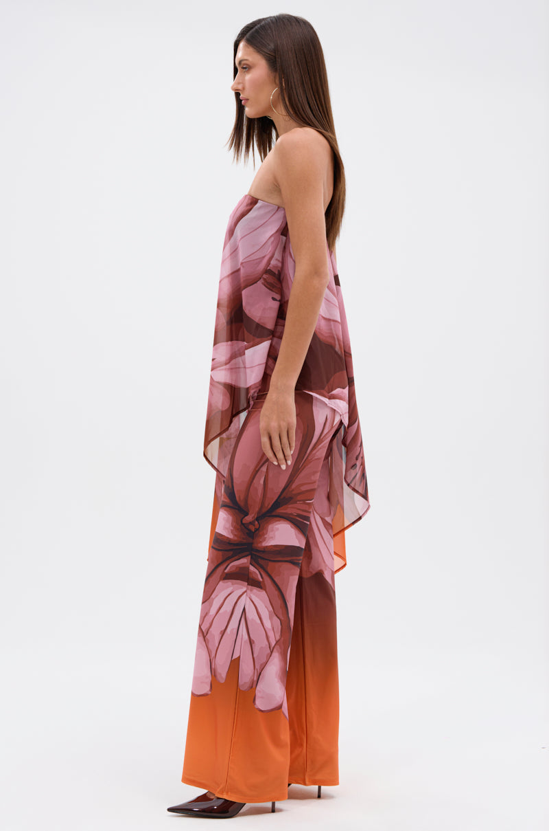 A woman stands in profile wearing the ON A BEACH SOMEWHERE PRINTED JUMPSUIT IN ORANGE—strapless, flowy, and bold with a tropical floral pattern—perfect for summer parties, paired with pointed brown shoes against a plain white background. 2