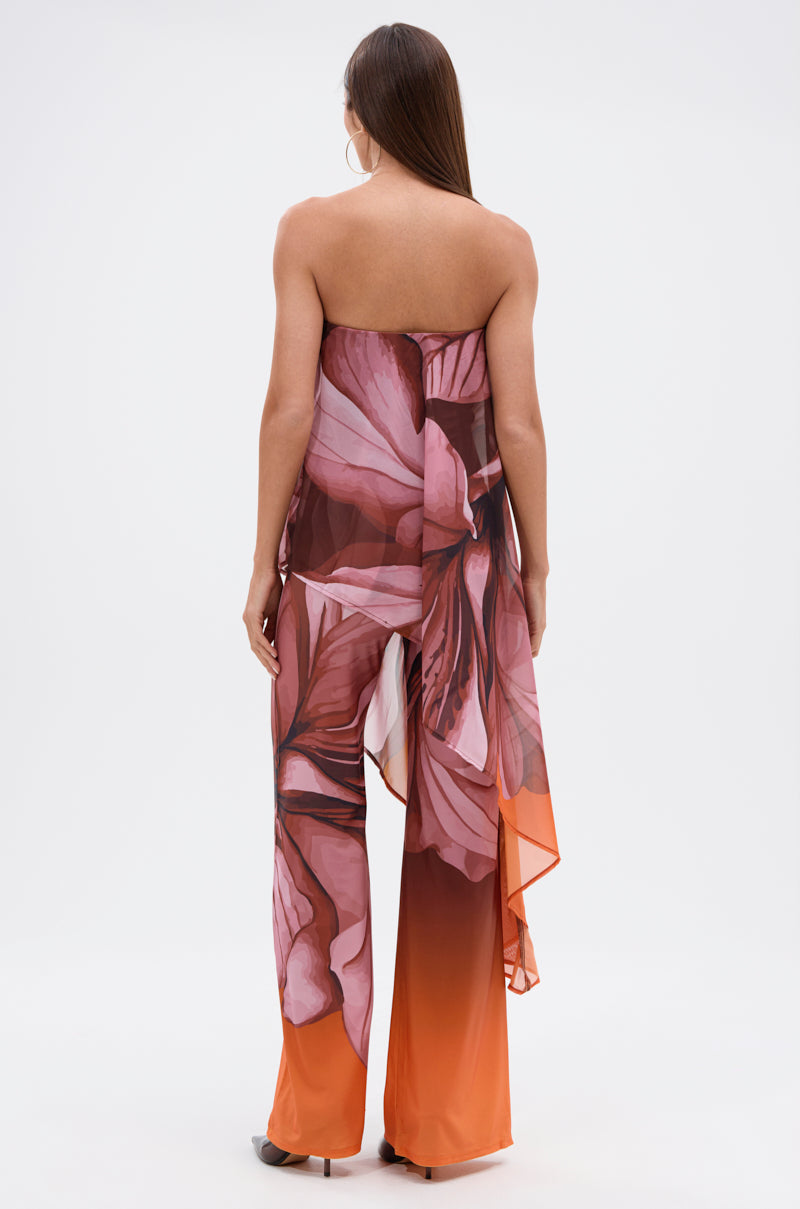 A woman with long brown hair wears the ON A BEACH SOMEWHERE PRINTED JUMPSUIT IN ORANGE, a strapless, flowy jumpsuit with a tropical floral print and asymmetrical hem, standing against a plain white background, photographed from behind. 3