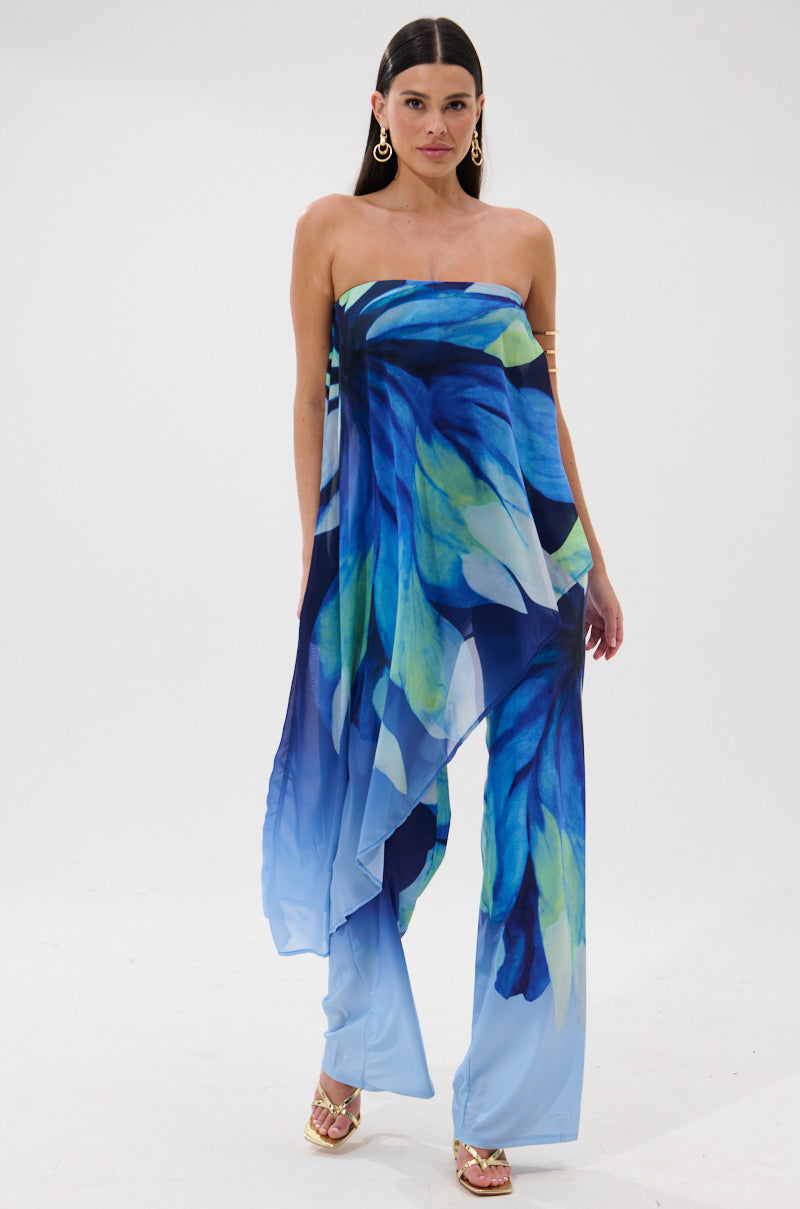 A woman models the ISLAND QUEEN JUMPSUIT, a strapless tropical print piece with blue, green, and aqua abstract leaves, flared legs, gold hoop earrings, and gold high-heeled sandals against a white background.