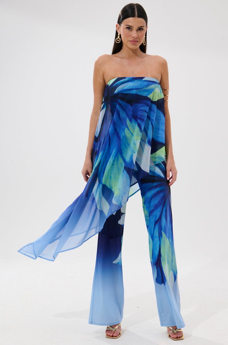 A woman wears the ISLAND QUEEN JUMPSUIT, a strapless, flowing jumpsuit with a blue, green, and yellow abstract leaf print and flared legs. She pairs it with gold hoop earrings and open-toe heels, standing against a white background.