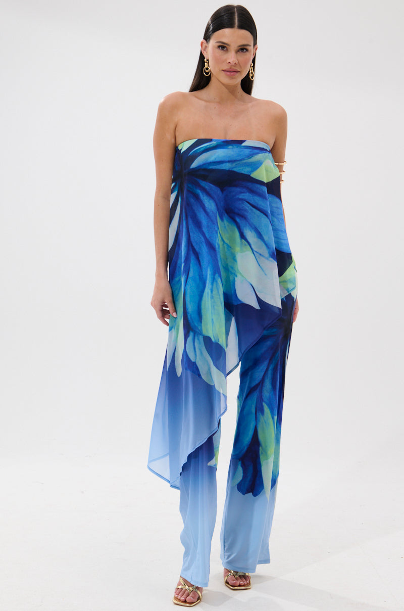 A woman with long dark hair, gold earrings, and strappy heels models the ISLAND QUEEN JUMPSUIT—a strapless, flowy vacation piece featuring a bold blue, green, and white tropical petal print—against a white background.