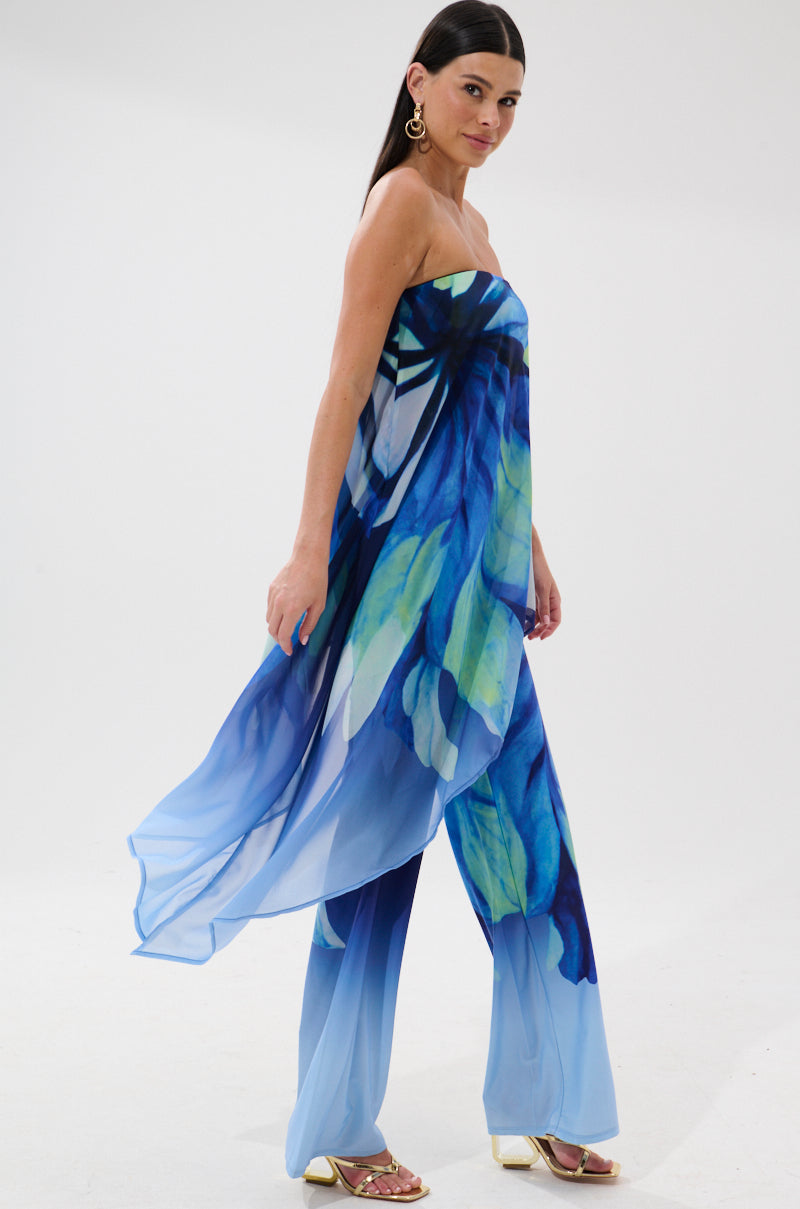 Confident woman in the ISLAND QUEEN JUMPSUIT, a strapless, flowing blue tropical print with bold florals, styled with gold earrings and sandals, walking against a plain white background.