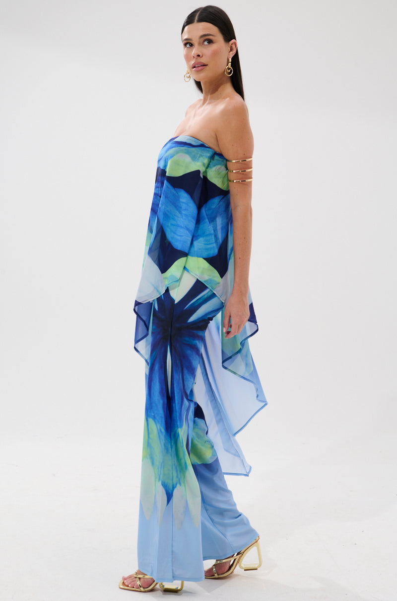 Model in the ISLAND QUEEN JUMPSUIT—a strapless blue and green tropical print jumpsuit with a flowing asymmetrical overlay—styled with gold arm cuff, hoop earrings, and gold heels. Perfect vacation look against a white background.